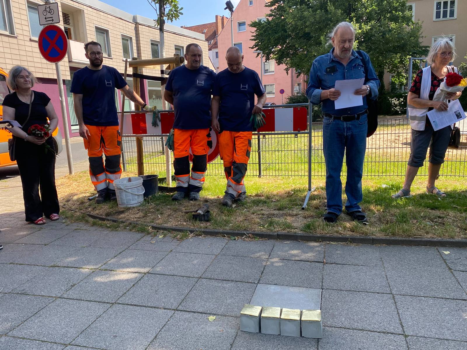 Vier quadratische Steine liegen auf einem Fu&szlig;weg, dahinter stehen Menschen: Au&szlig;en je eine Frau mit Blumenstrau&szlig;, drei M&auml;nner mit oranger Arbeitshose und blauem T-Shirt und ein Mann in Jeans, der eine Seite Papier h&auml;lt und darauf schaut.