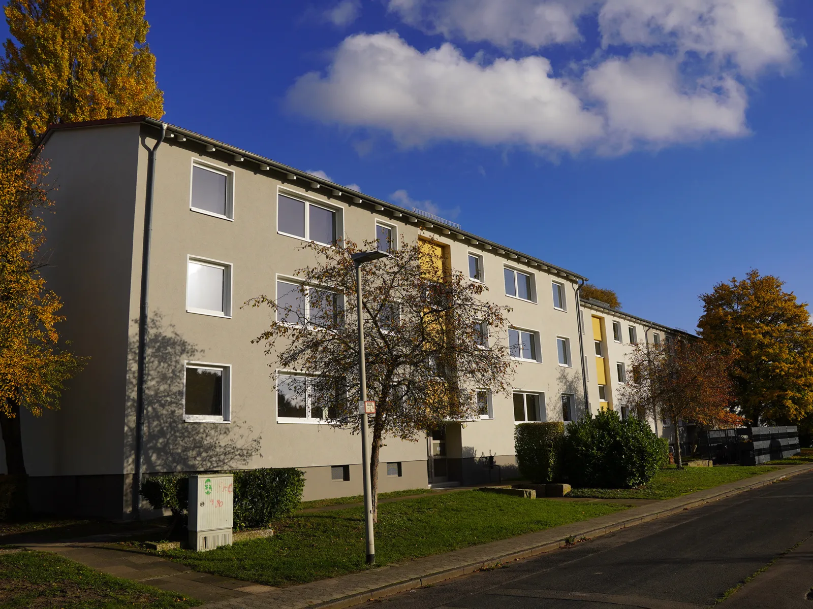 Ein langes Wohngebäude mit zwei Obergeschossen, davor Rasen und ein Baum, blauer Himmel mit weißen Wolken.