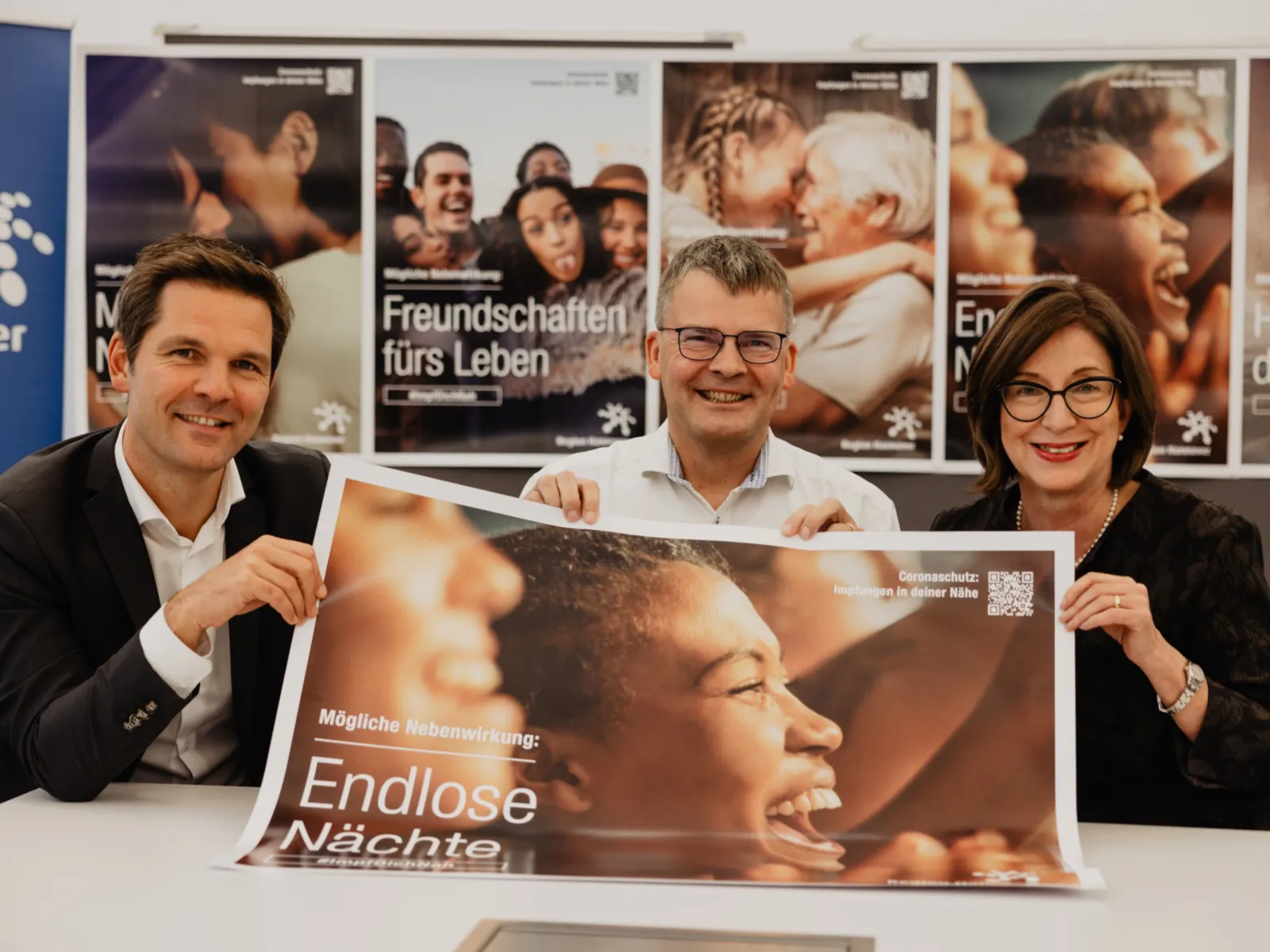 Zwei Männer und eine Frau sitzen an einem Tisch und halten gemeinsam ein Plakat.