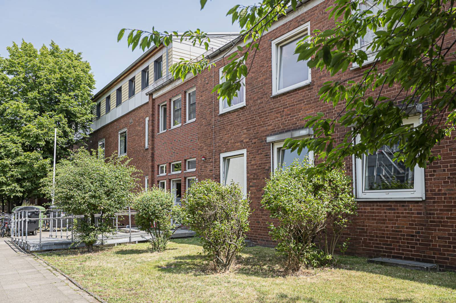 Ein Gebäude aus roten Backsteinen. Eine flache Rampe führt zum Eingang. Vor dem Gebäude ein Grünstreifen mit einigen Büschen.