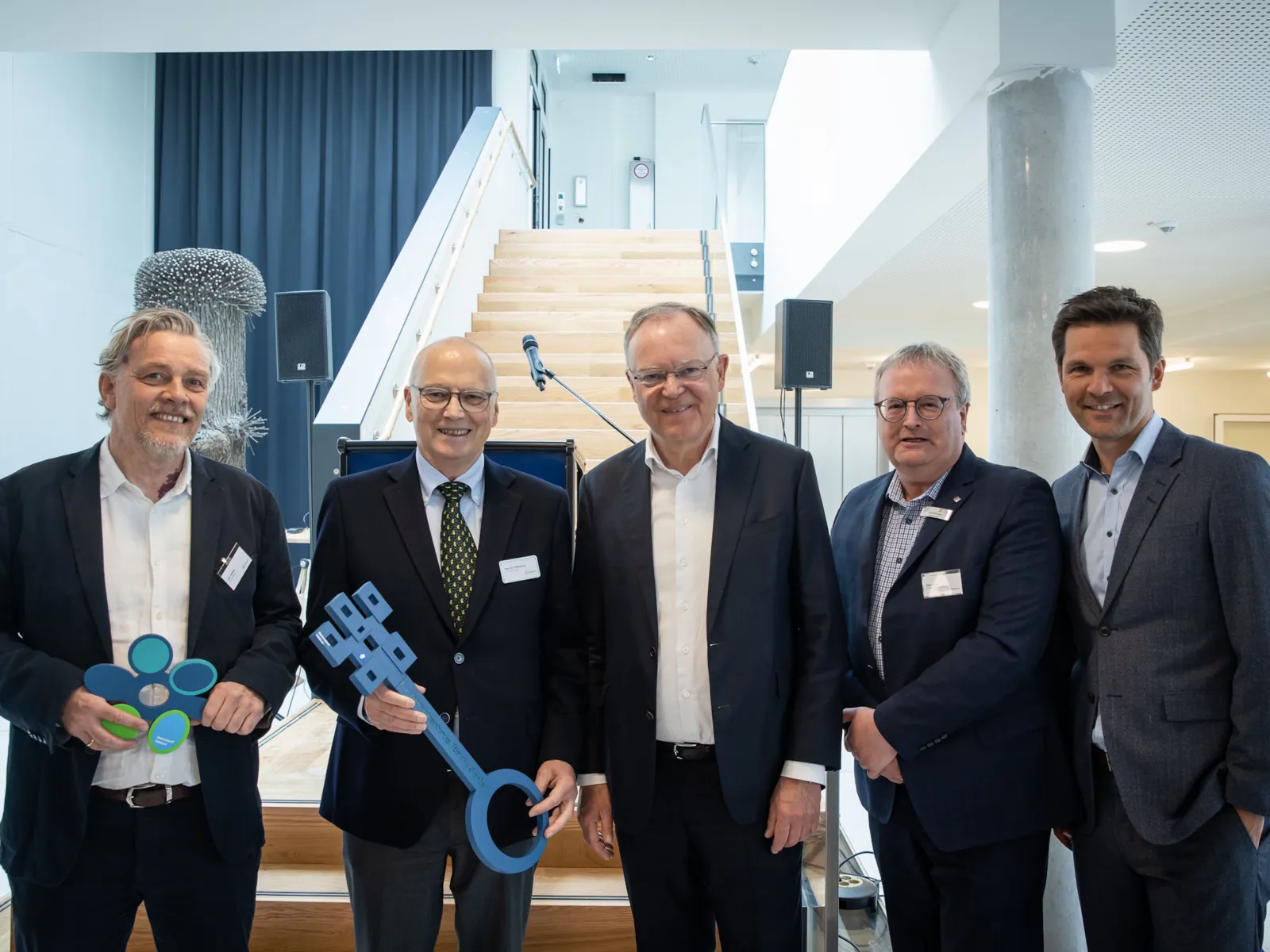 Fünf Männer in Anzügen, einer hält einen übergroßen symbolischen Schlüssel in der Hand.