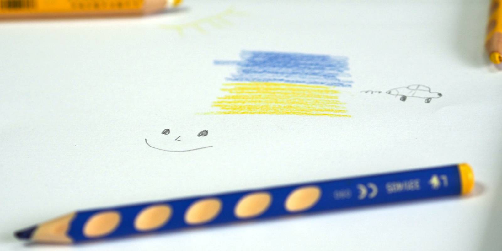 Einfache Zeichnung mit Buntstiften auf einem weißen Blatt Papier: Die Farben Blau und Gelb, eine Sonne, ein grinsendes Gesicht und ein Auto. Auf dem Blatt verteilt liegen Buntstifte.