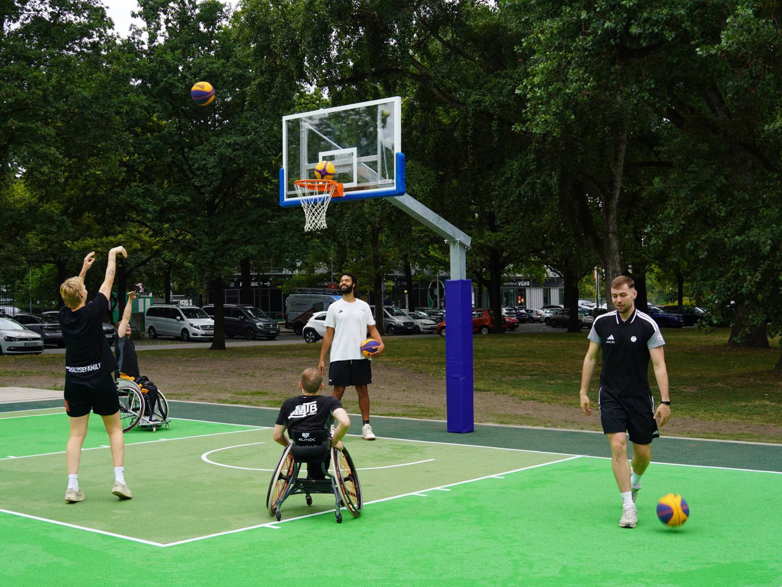 F&uuml;nf M&auml;nner in Sportkleidung auf einem gr&uuml;nen Feld, einer wirft einen Ball in einen Basketballkorb. Drei M&auml;nner stehen, zwei sitzen in Sport-Rollst&uuml;hlen.