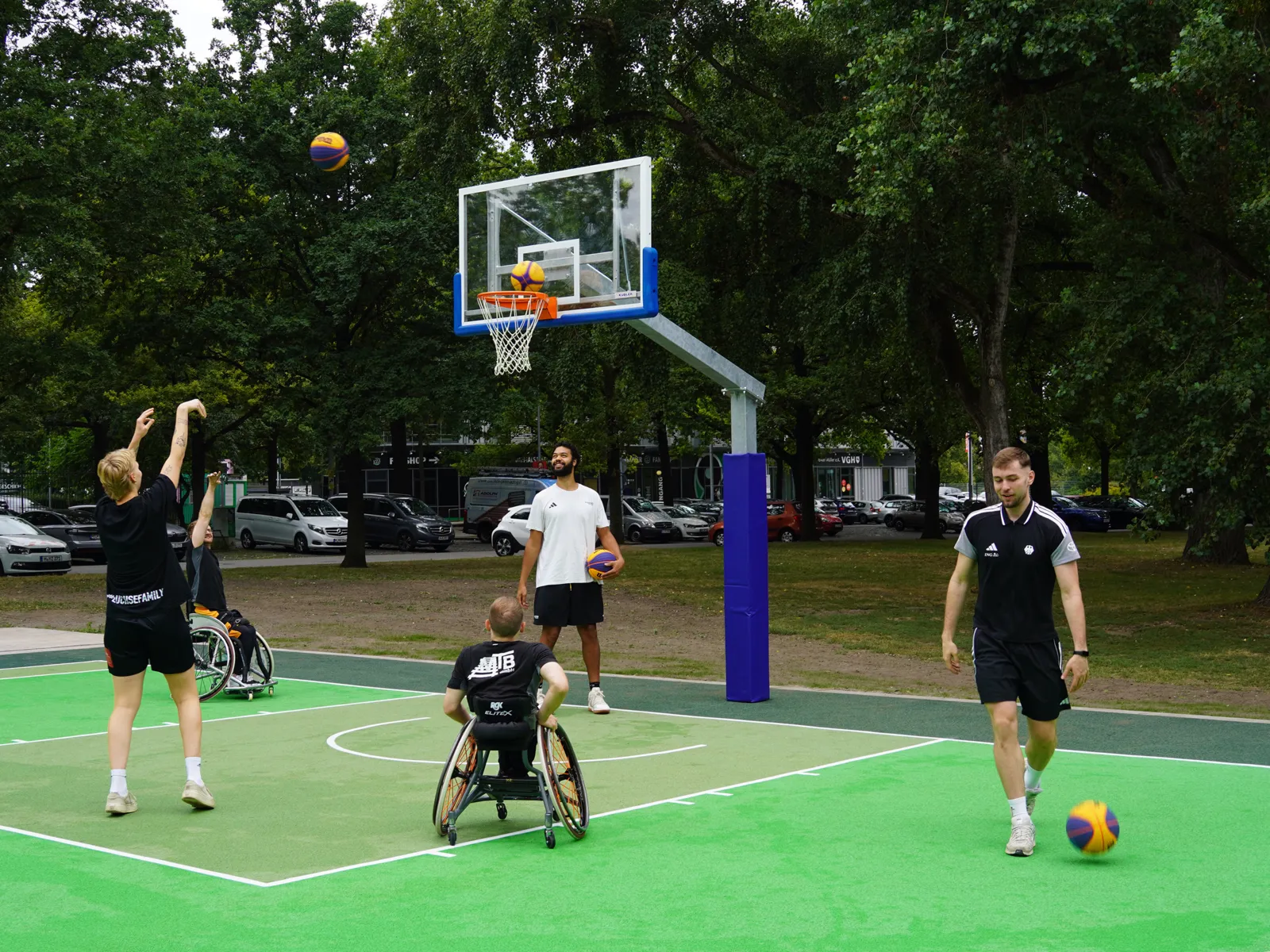 F&uuml;nf M&auml;nner in Sportkleidung auf einem gr&uuml;nen Feld, einer wirft einen Ball in einen Basketballkorb. Drei M&auml;nner stehen, zwei sitzen in Sport-Rollst&uuml;hlen.