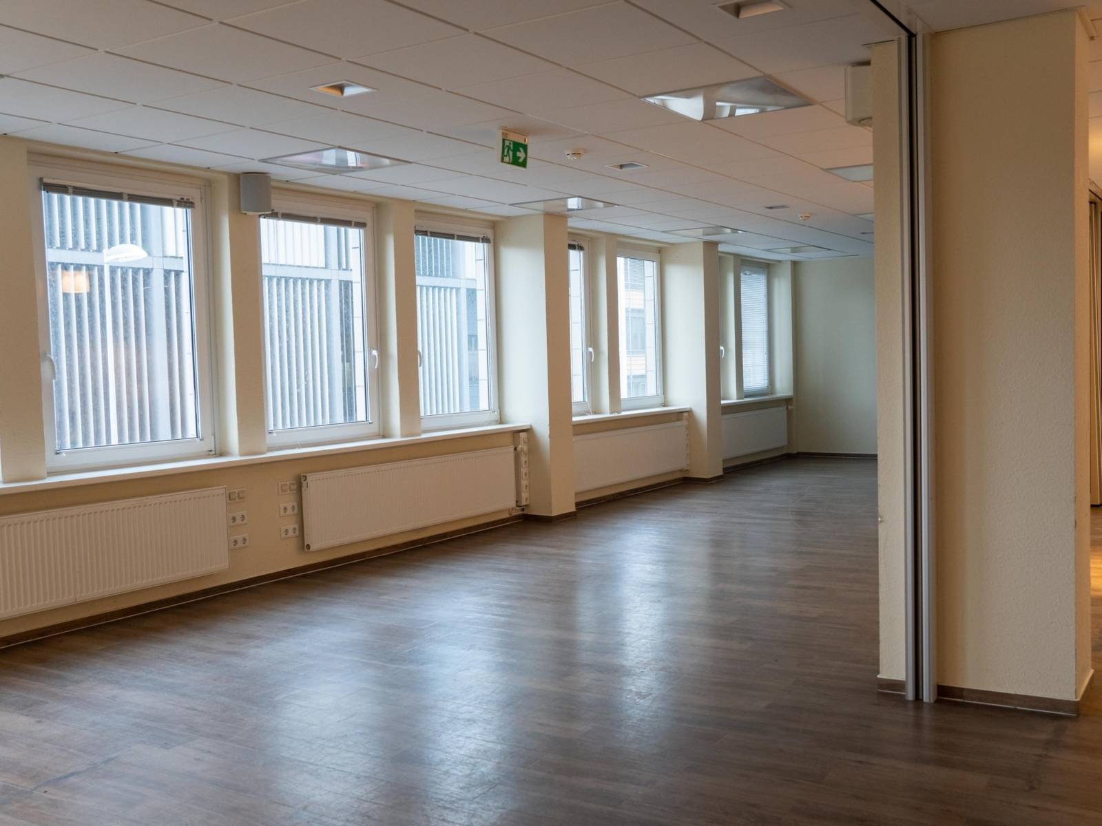 Ein leerer Raum mit Holzfu&szlig;boden, einer Fensterfront und Heizungen unter den Fenstern.