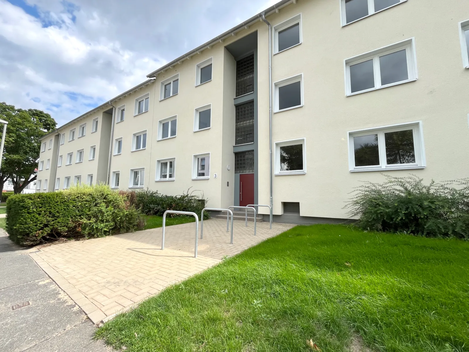 Dreigeschossige Wohnhäuser mit gelber Fassade, davor Fahrradbügel auf den Zuwegungen, daneben grüner Rasen und eine Hecke.
