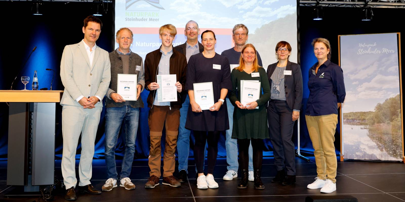Neun Personen stehen auf einer Bühne, dabei werden vier Urkunden gehalten.