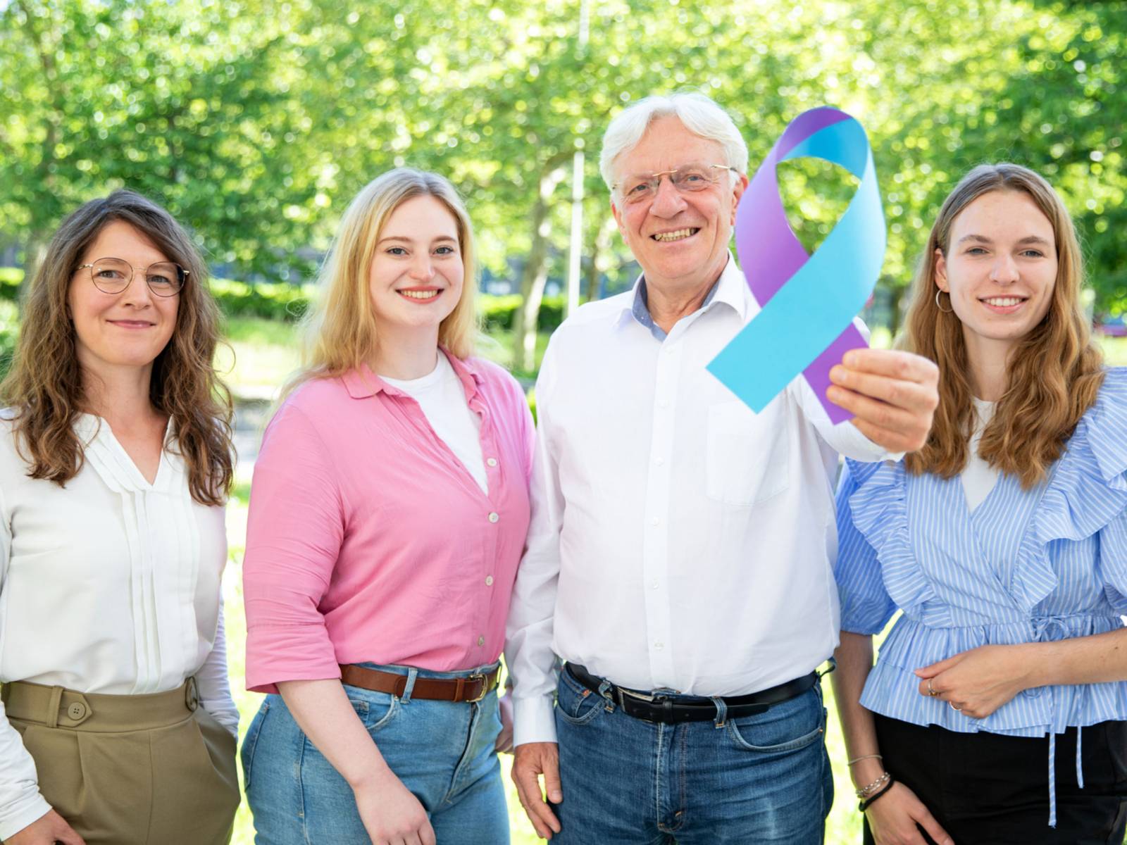 Drei Frauen und ein Mann halten eine blau-lila Schleife in die Kamera.