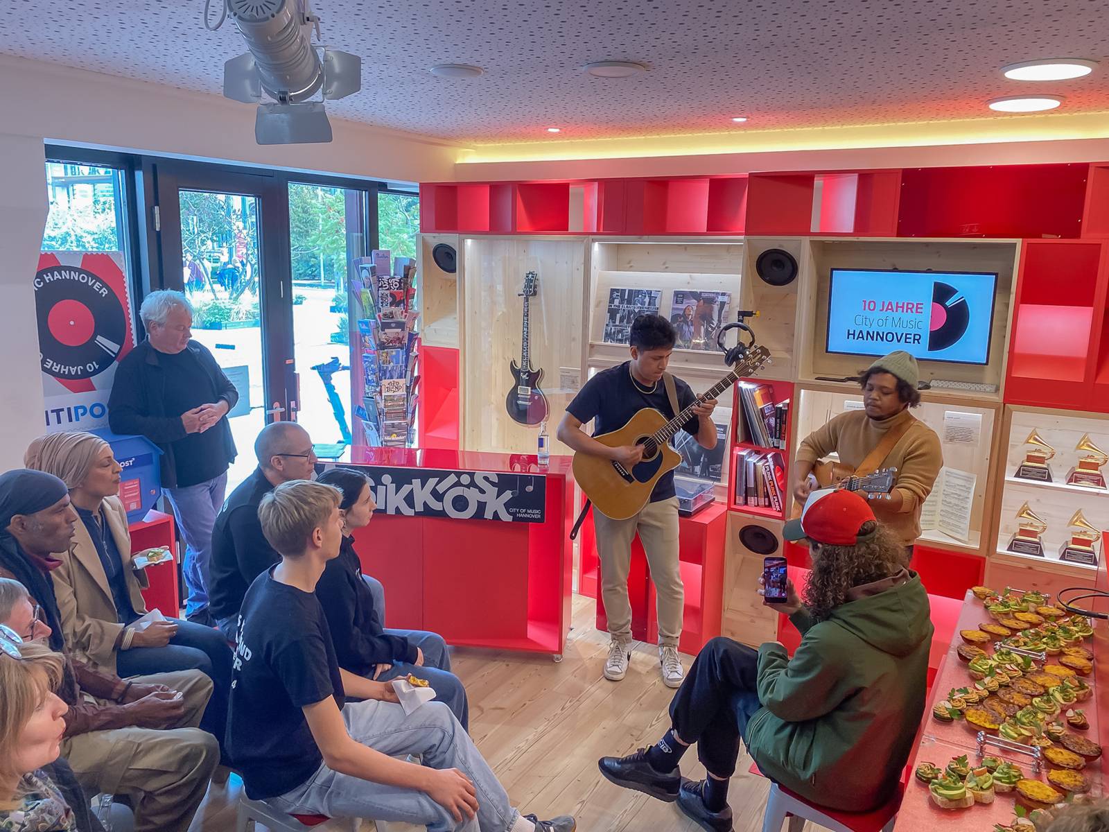 In einem kleinen Raum stehen zwei Männer mit Gitarren und musizieren, drum herum sitzt ein kleines Publikum.