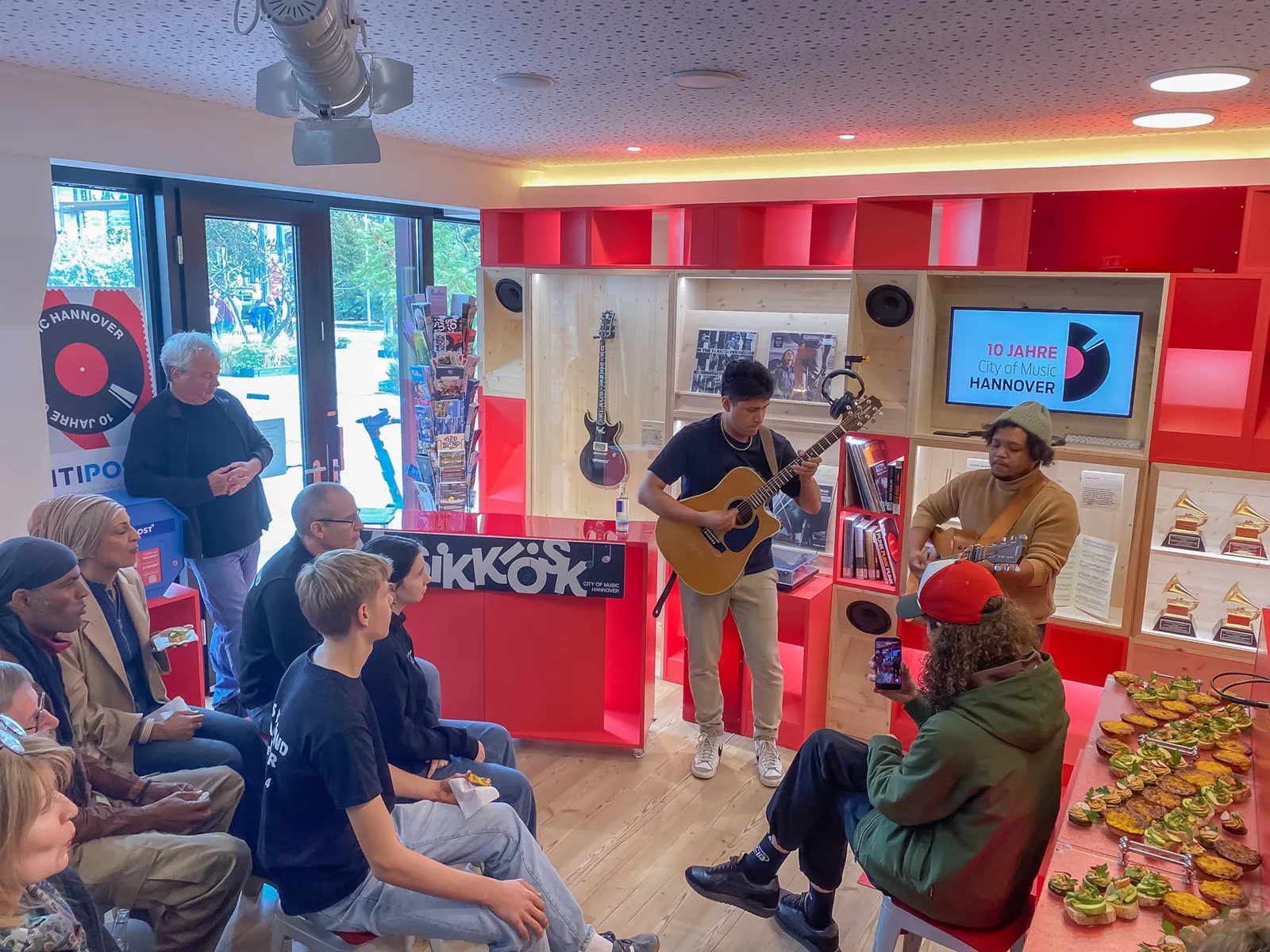 In einem kleinen Raum stehen zwei M&auml;nner mit Gitarren und musizieren, drum herum sitzt ein kleines Publikum.