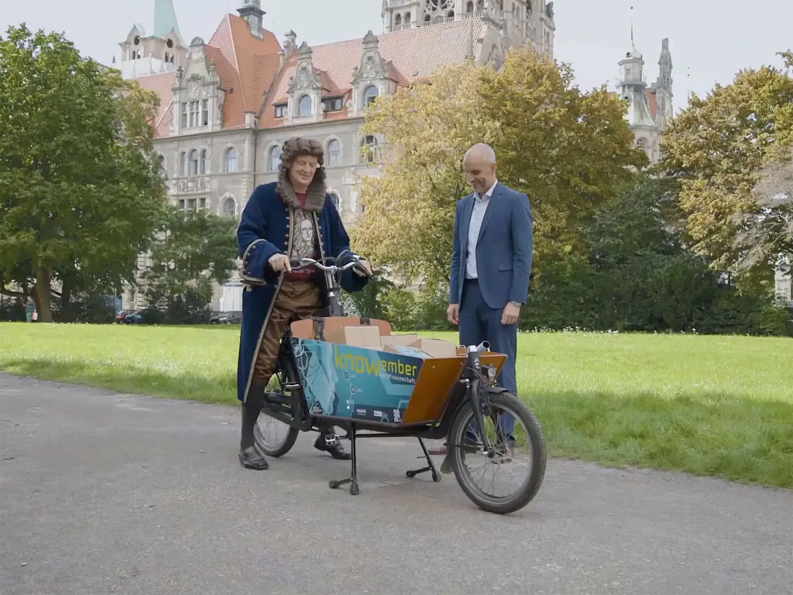 Ein Mann mit Perücke in Kleidung aus der Barockzeit auf einem Lastenrad und ein Mann im Anzug vor historischem Gebäude