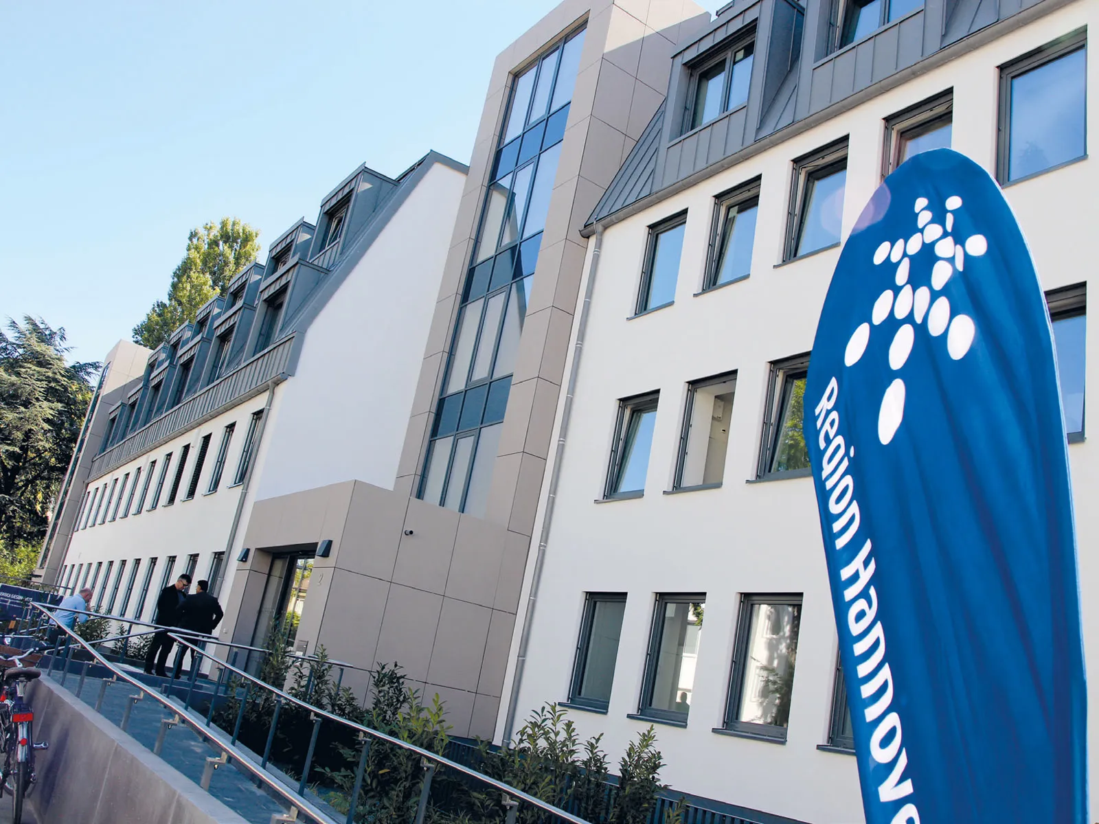 Ein modernes Gebäude mit einer Rampe für einen barrierefreien Zugang. Im Eingangsbereich stehen zwei Personen und eine weitere Person schließt ihr Fahrrad an. Vorne rechts im Bild ist ein blauer Aufsteller mit der Aufschrift "Region Hannover" zu sehen (das Wort Hannover ist zum Teil abgeschnitten).
