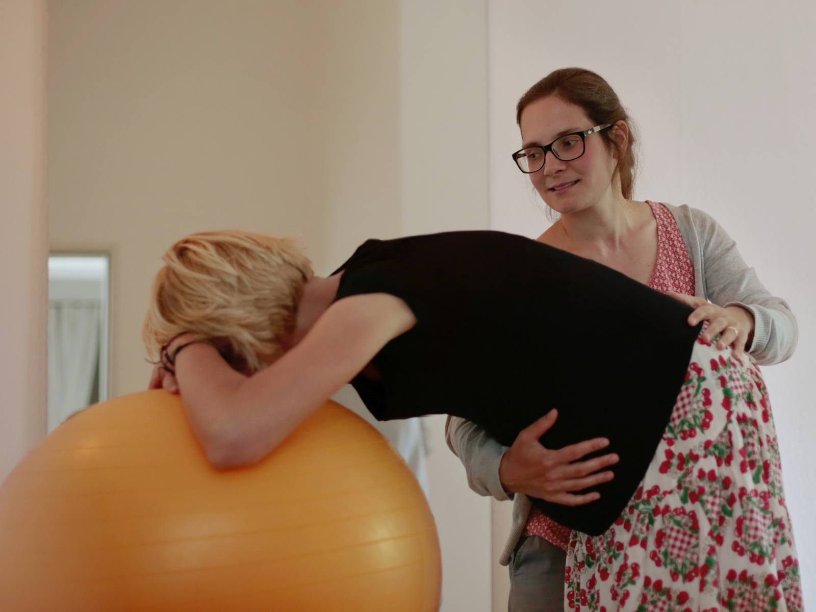 Zwei Frauen, von denen eine sich auf einem Gymnastikball aufstützt, während die andere sie hält