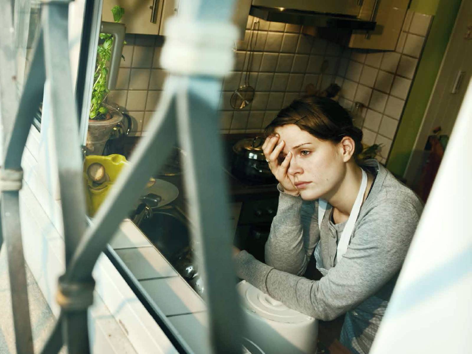 Eine Frau schaut mit leerem Blick aus einem Küchenfenster. Das Fenster ist von außen vergittert.
