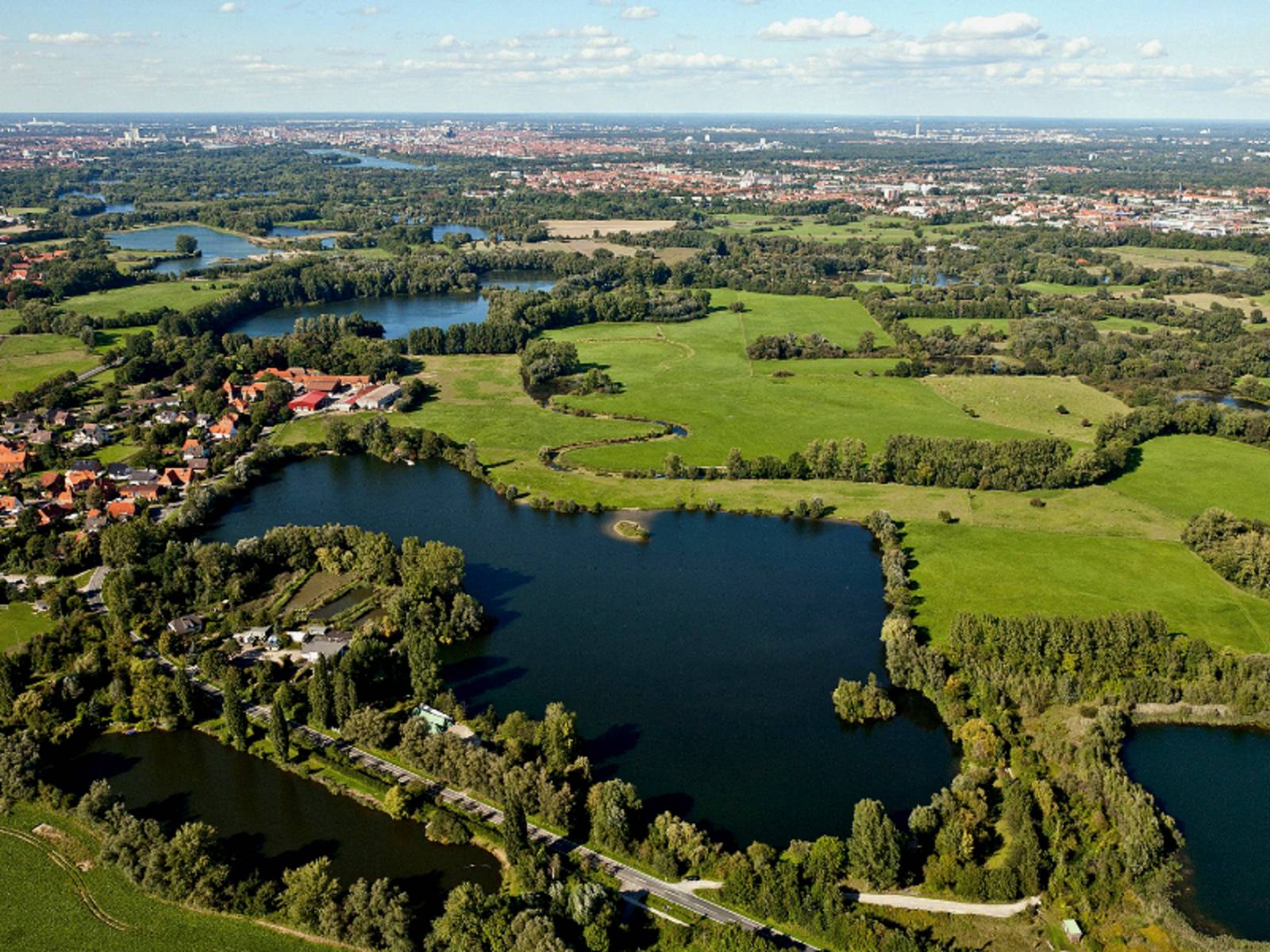 Luftaufnahme: Mehrere kleine Gewässer sind durch Gräben miteinander verbunden. Die Gewässer werden unterbrochen von Grünflcäehn, Bäumen und kleinen Siedlungen. Im Hintergrund ist die Stadt Hannover mit Maschsee und Fernsehturm zu erkennen.
