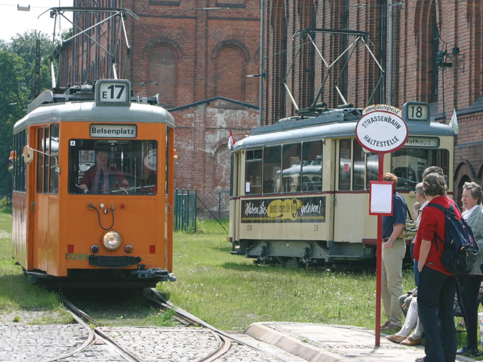 Zwei historische Straßenbahnen und Passanten an einer alten Straßenbahnhaltestelle