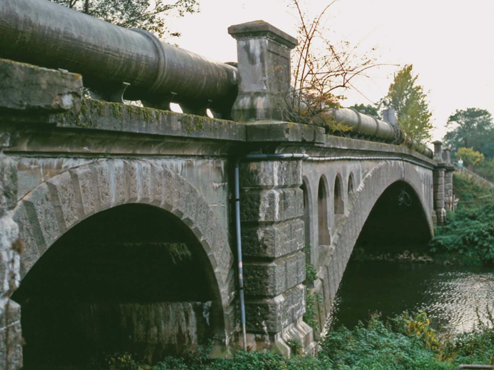 Ältere Brücke aus Stein über die ein sehr dickes Rohr läuft