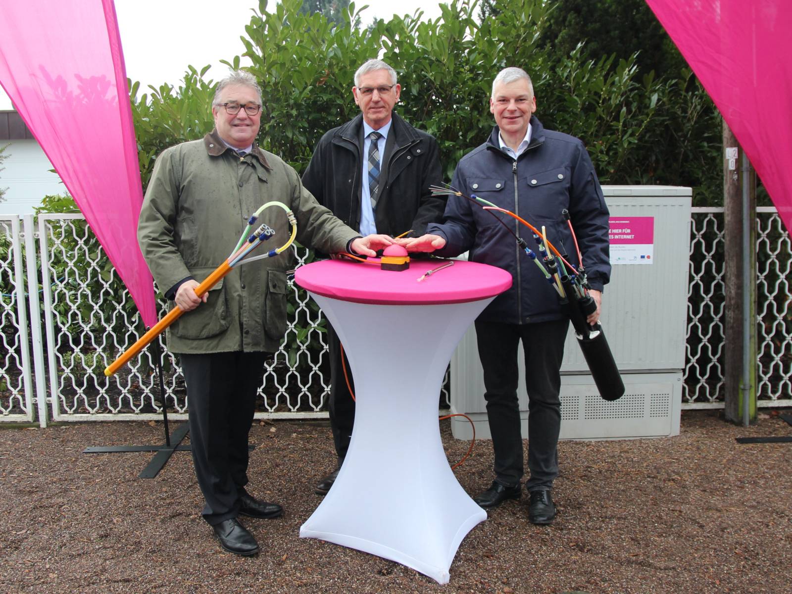 Drei Herren stehen draußen um einen Stehtisch mit pinkfarbener Tischplatte und drücken gemeinsam auf einen Buzzer.