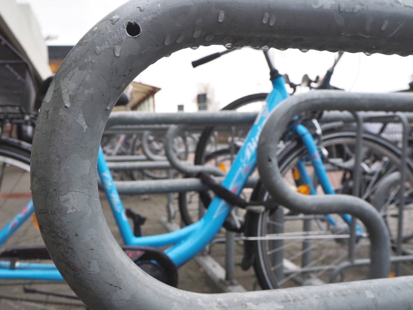 Blick auf einen Fahrradabstellbügel auf andere Bügel, an denen Fahrräder abgestellt sind. Regentriopfen haben sich an den Unterseiten des Metallbügels gesammelt.