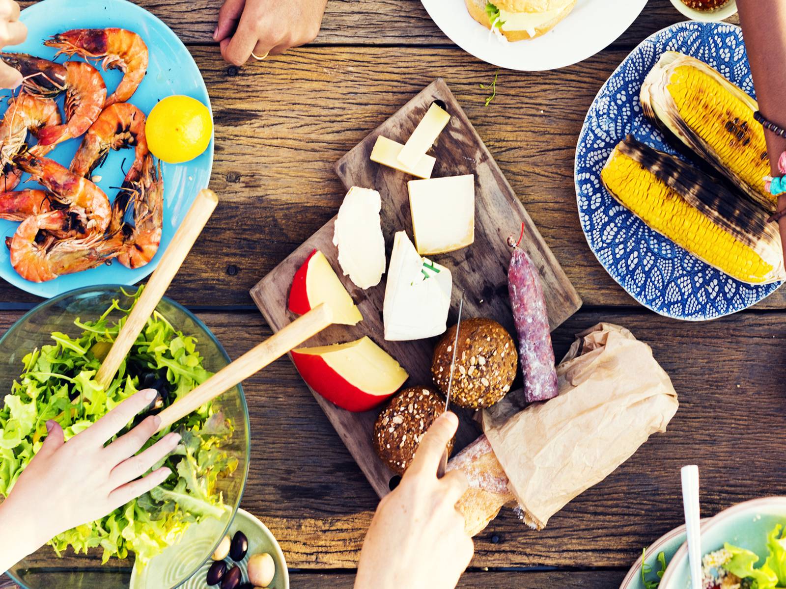 Holztisch auf dem Salat, Garnelen, Maiskolben, Wurst und Käse stehen. Hände von Personen, die sich bedienen