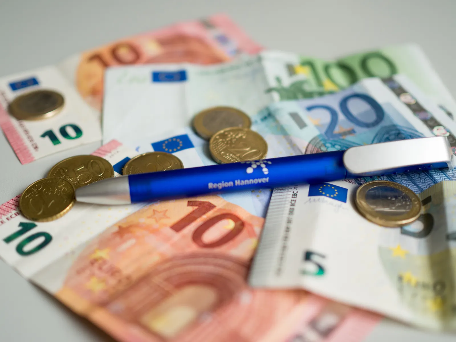 Zwei Zehneuroscheine, ein Hunderteuroschein, ein Zwanzigeuroschein, ein Fünfeuroschein, mehrere Münzen und ein blauer Kugelschreiber liegen auf einem grauen Tisch.