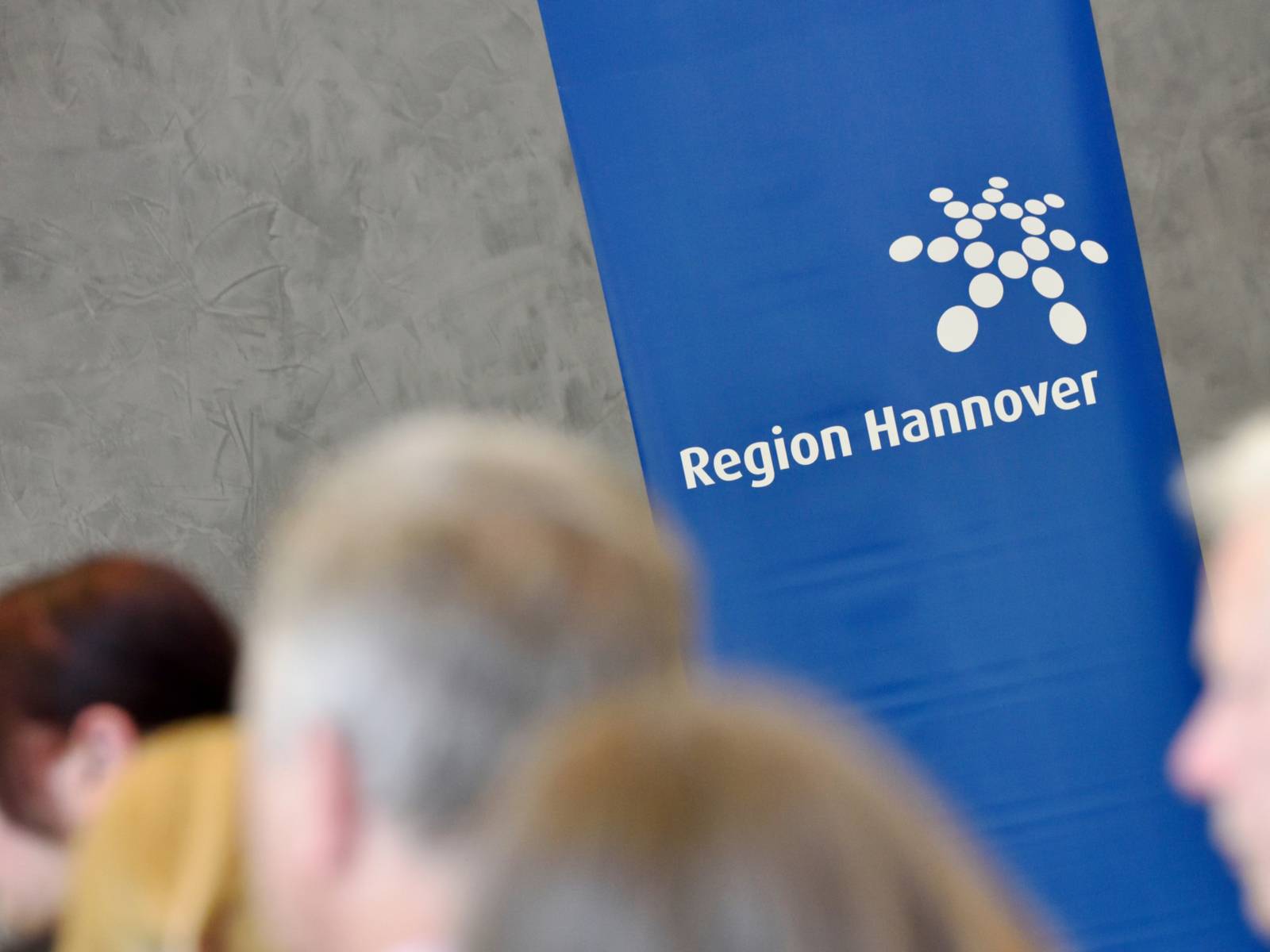 Auf einem Banner ist das Logo der Region Hannover in Weiß auf blauem Grund zu sehen. Drumherum stehen Menschen.