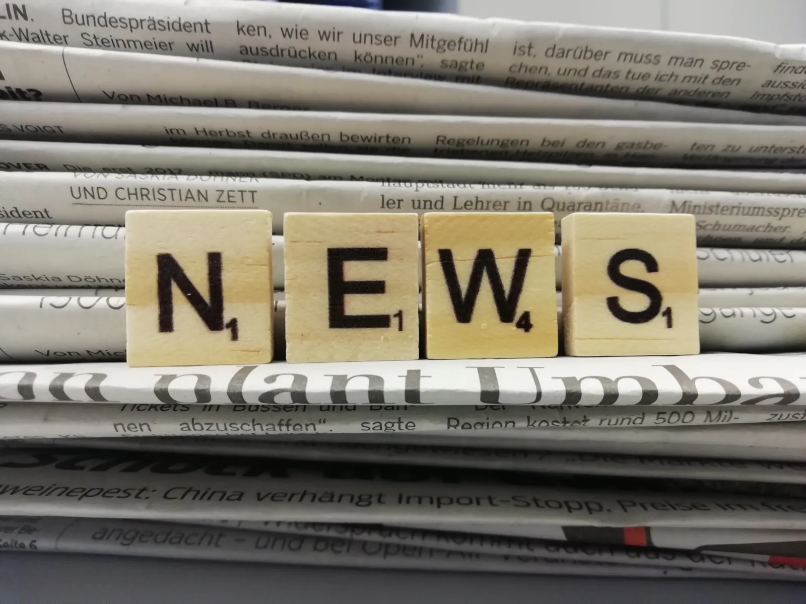 Vier Holzwürfel mit Buchstaben drauf, die nebeneinander aufgestellt das Wort "News" ergeben. Dahinter ist ein Stapel Zeitungen zu sehen.