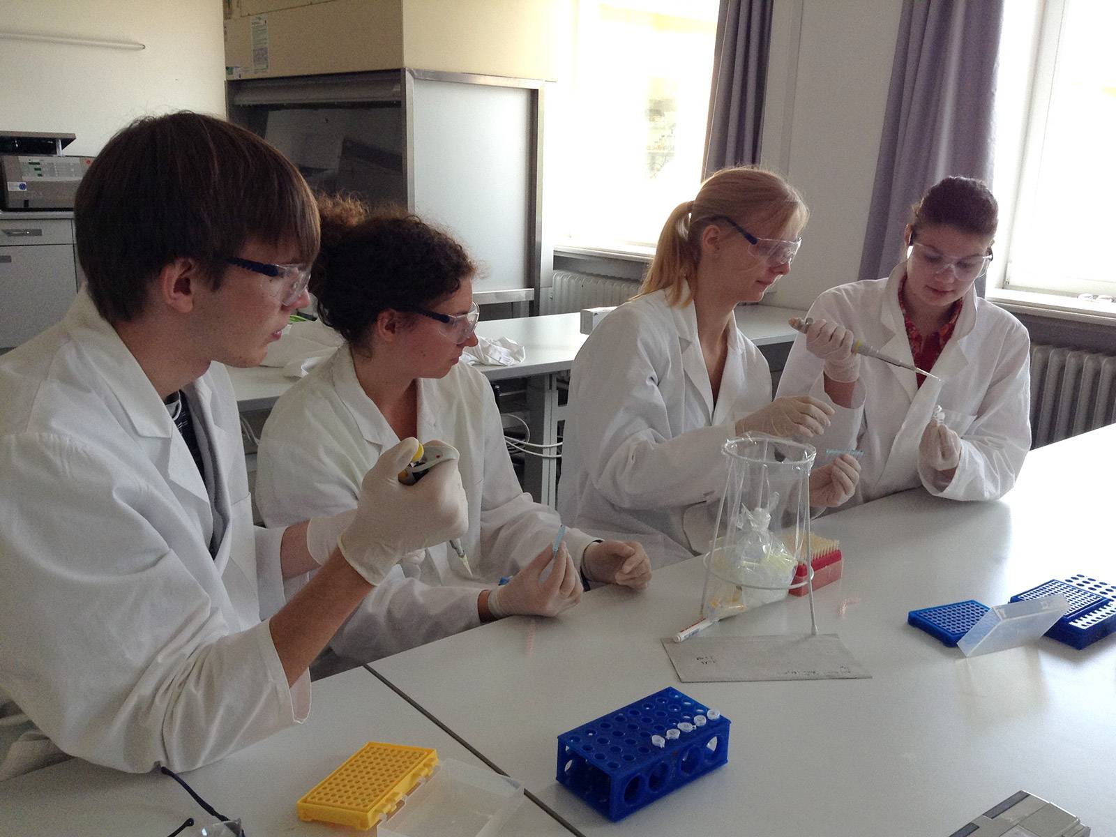 Ein Schüler und drei Schülerinnen in weißen Kitteln und mit Schutzbrillen arbeiten mit Pipetten in einer Laborumgebung