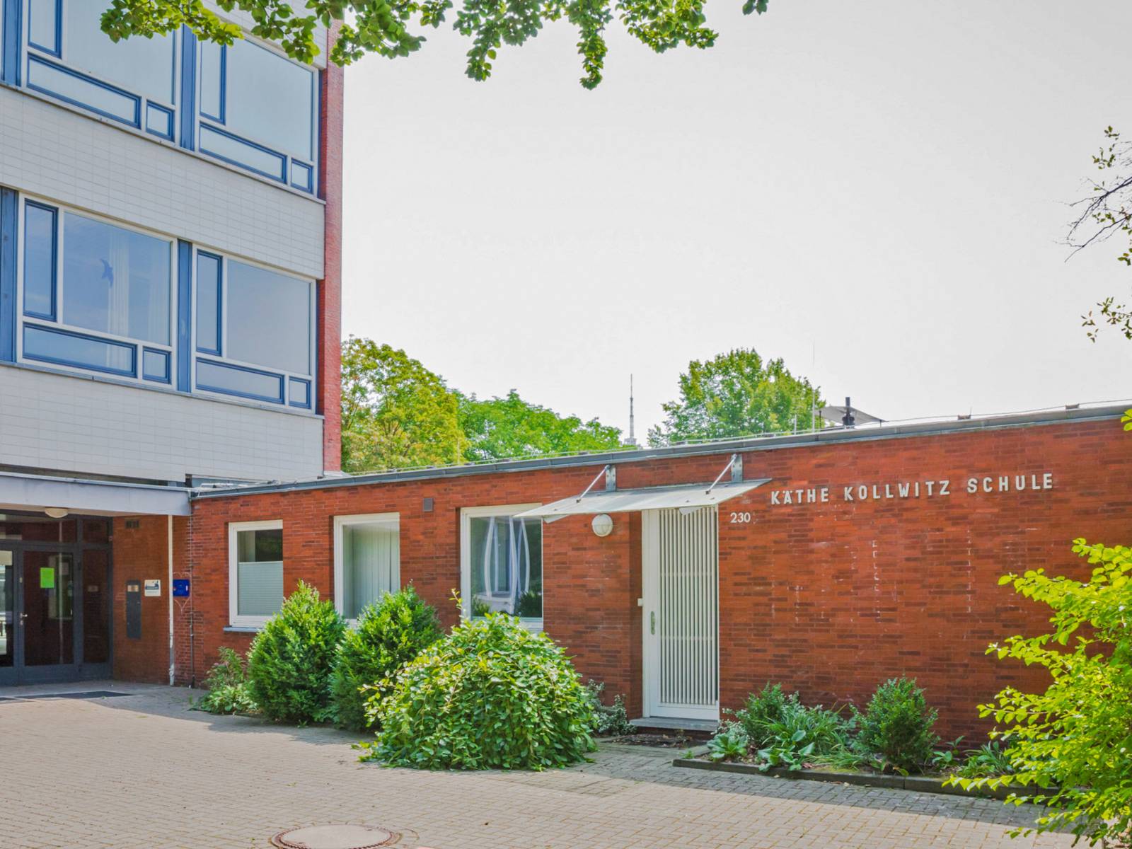 An einem roten einstöckigen Flachbau aus Backsteinen ist der Schriftzug "Käthe-Kollwitz-Schule" angebracht; daneben der Haupteingang zur Schule in einem mehrstöckigen Gebäude.