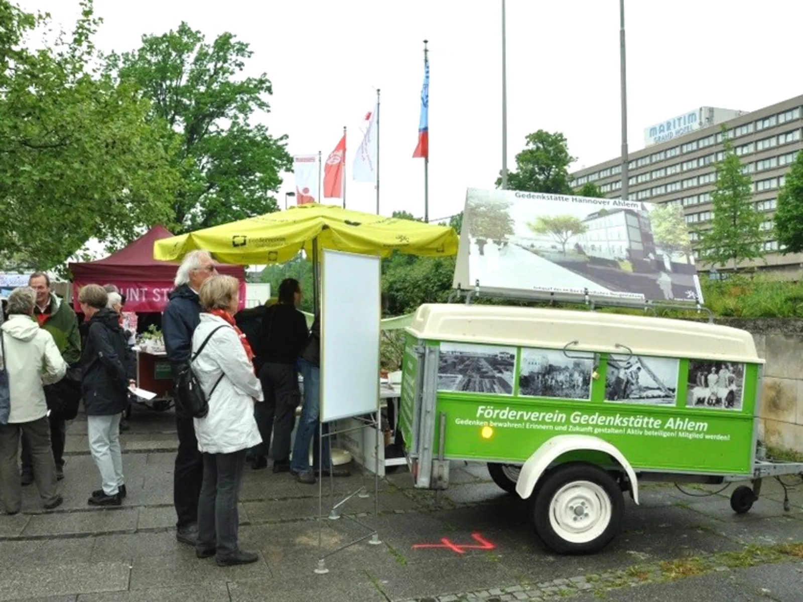Informationsstand des Fördervereins Gedenkstätte Ahlem an dem BesucherInnen stehen. Ein grüner Werbeanhänger des Fördervereins steht neben dem Infostand.