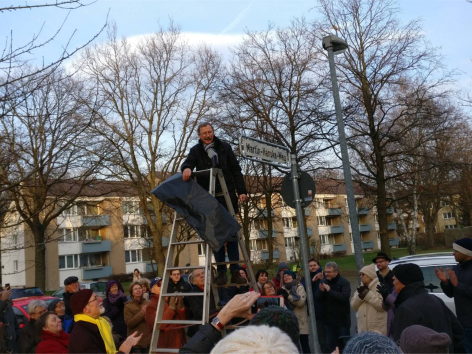 Ein Mann steht auf einer Leiter und enthüllt ein Straßenschild. Dabei stehen mehrere Personen, die dabei zusehen.