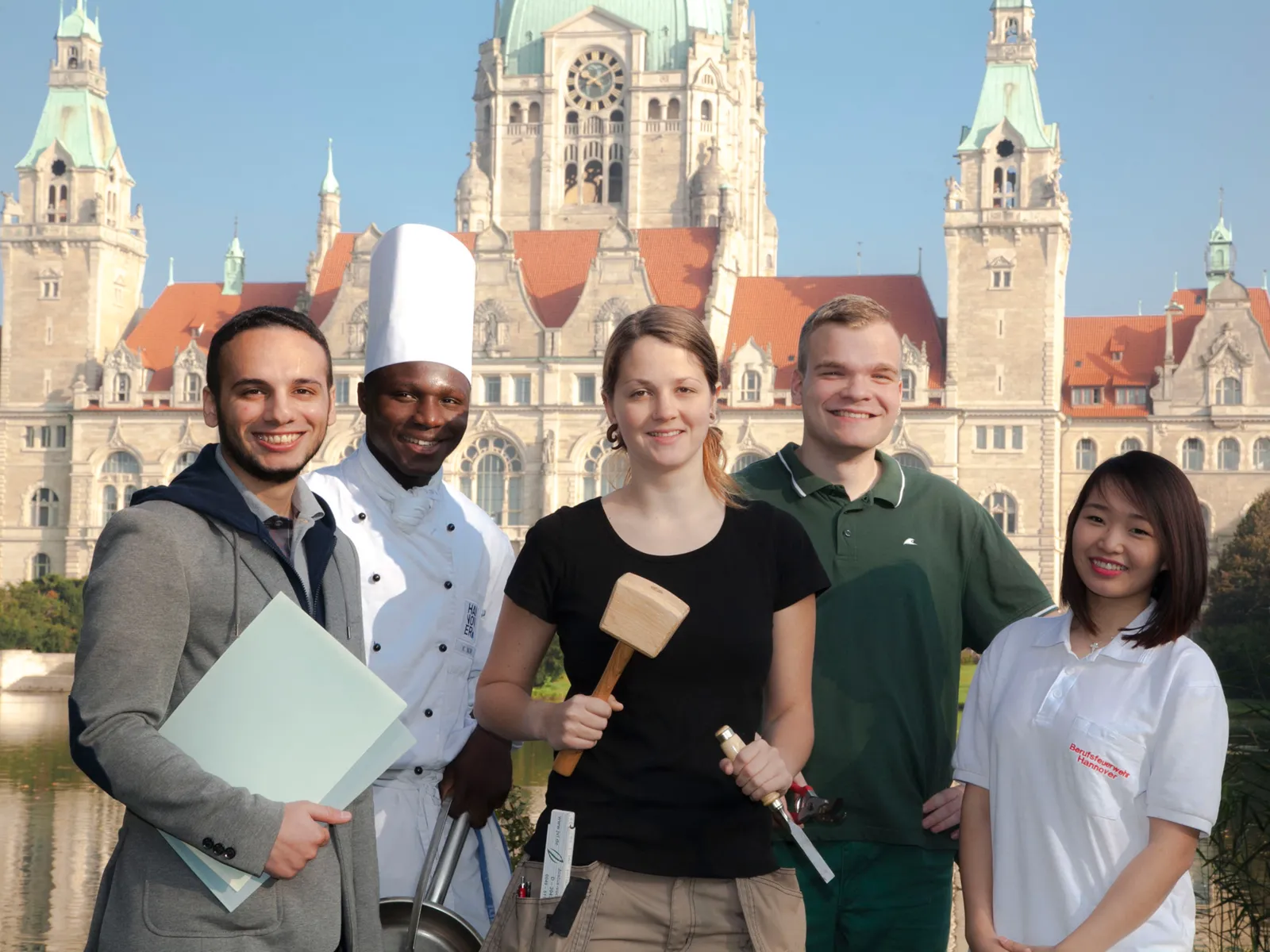 Azubis (drei Männer, zwei Frauen) verschiedener Berufsgruppen stehen zusammen vor dem Hannoverschen Rathaus - zum Teil mit Arbeitsutensilien