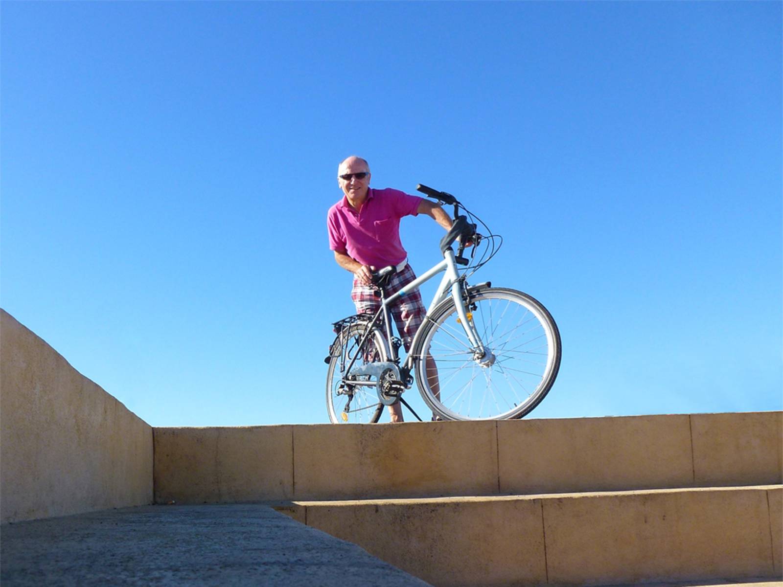 Älterer Herr steht mit Fahrrad vor blauem Himmel an einem Sommertag