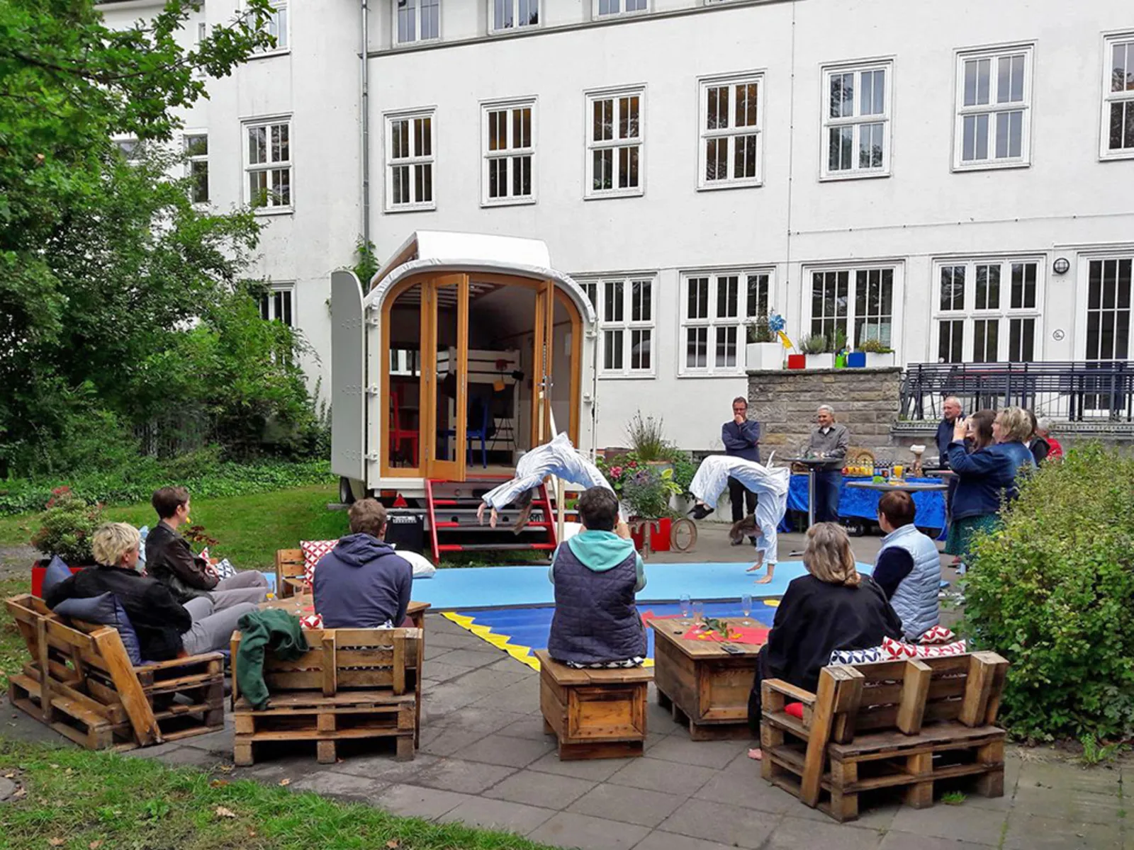 Vor einem geöffneten Zirkuswagen turnen zwei Akrobatinnen auf einer Matte. Die im Kreis darum sitzenden Besucherinnen und Besucher schauen zu.
