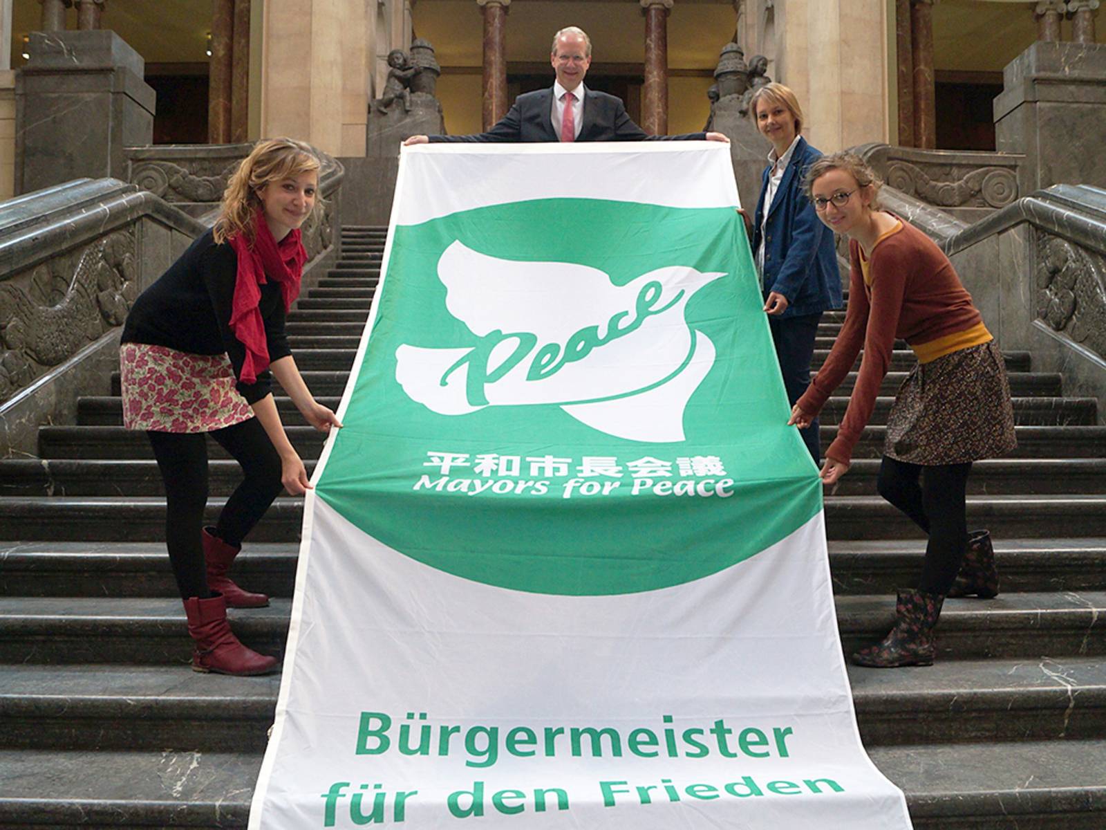 Vier Personen präsentieren auf der Treppe in der Rathaushalle die grün-weiße Flagge der Mayors for Peace