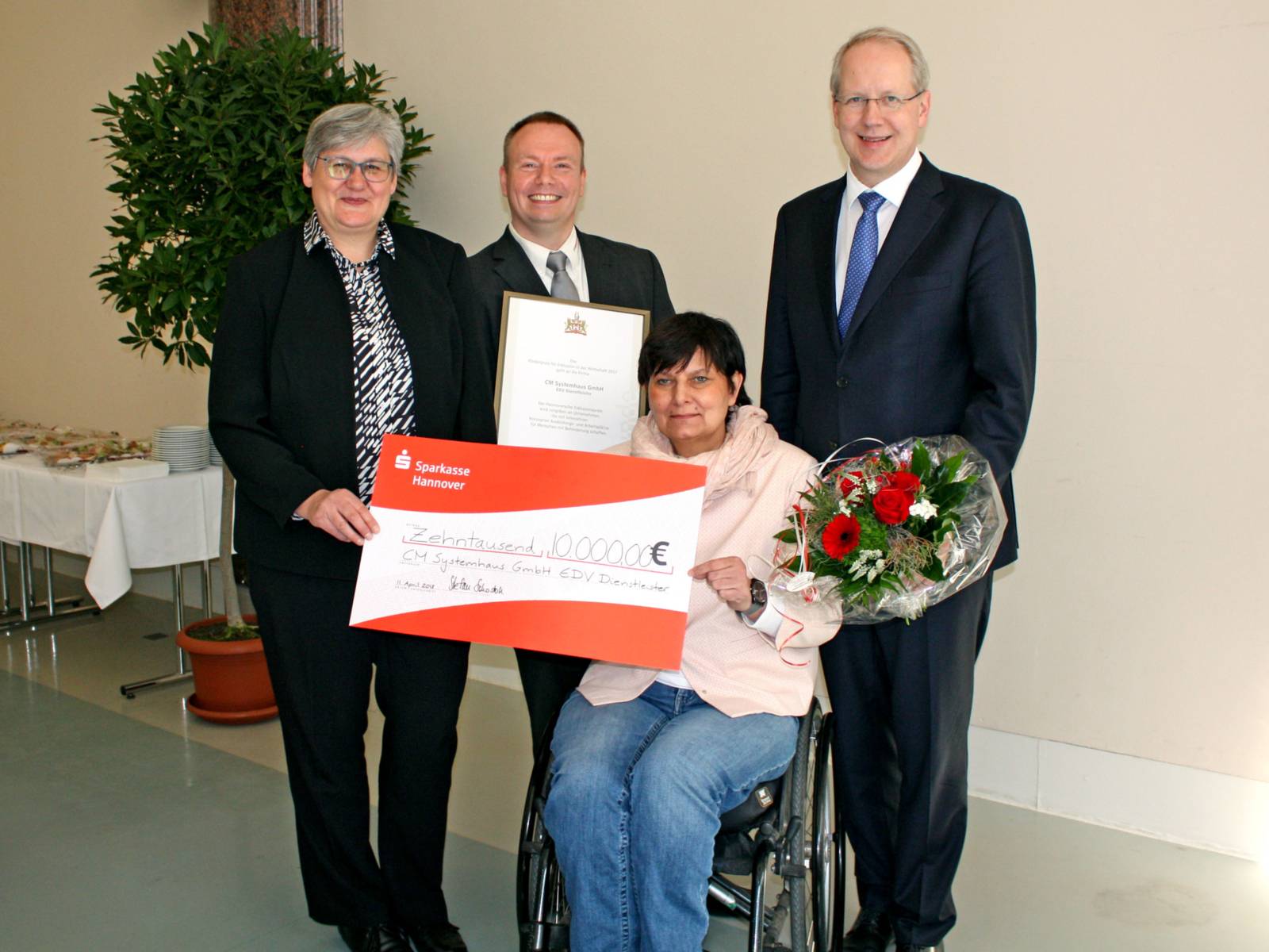 Zwei Frauen und zwei Männer, die gemeinsam eine Urkunde und einen großen Scheck halten. 