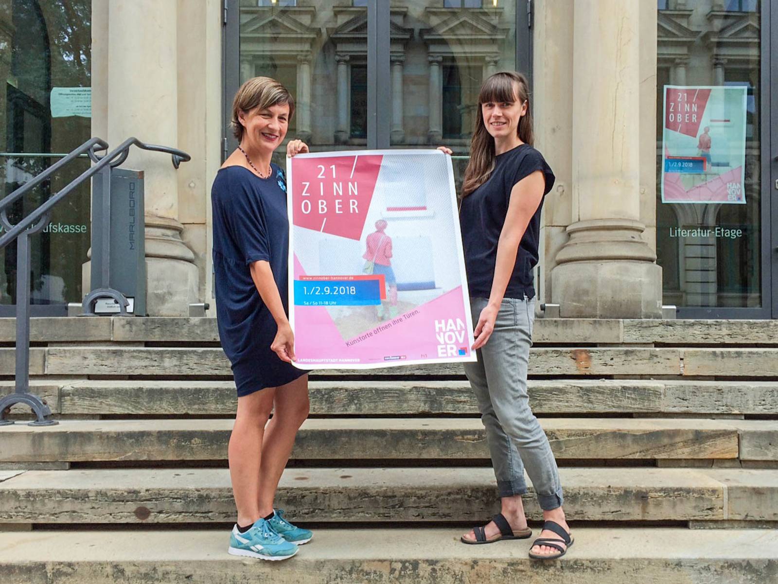 Zwei Frauen stehen auf einer Treppe und präsentieren das Plakat einer Kulturveranstaltung