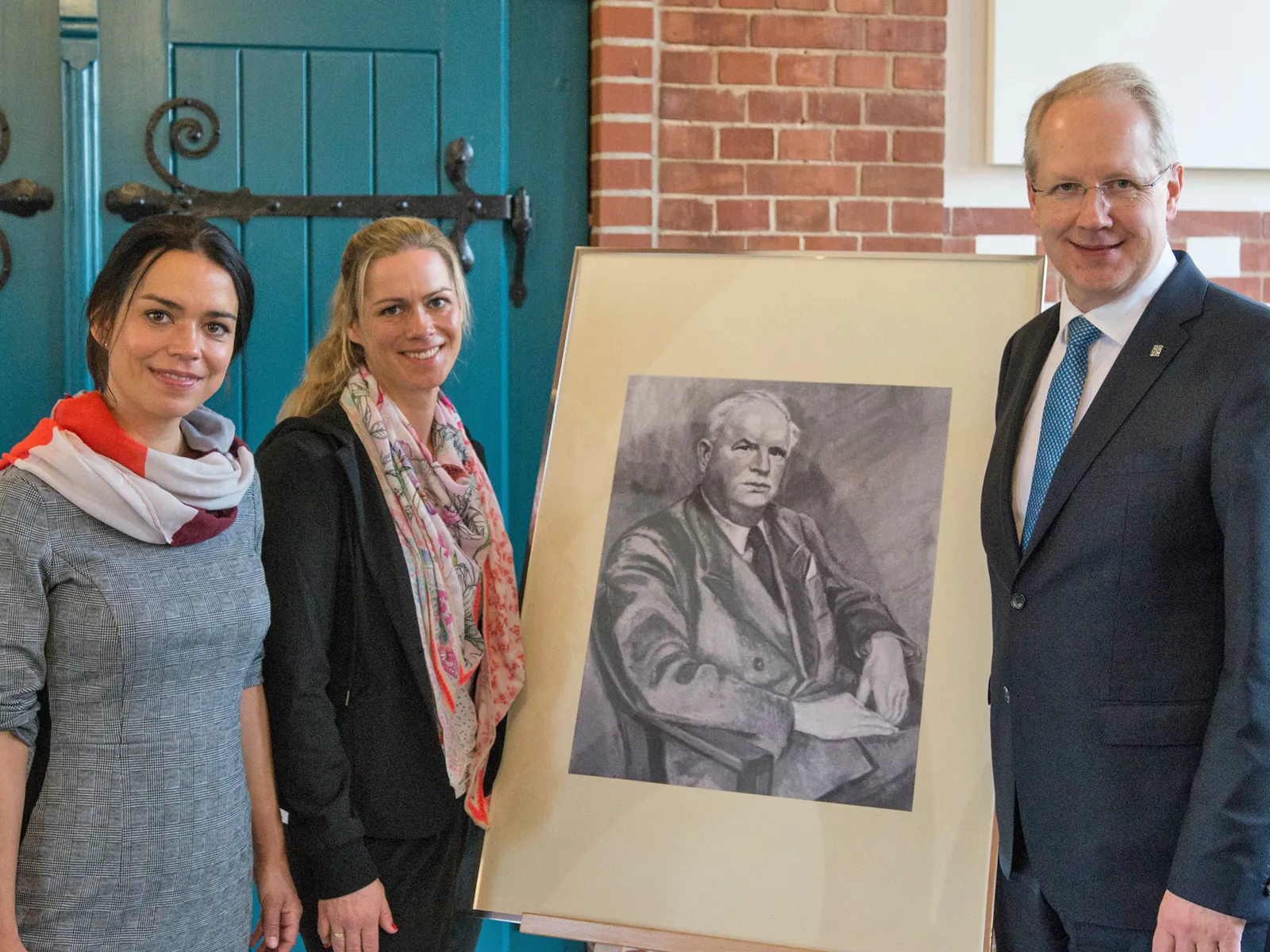 Drei Personen neben einer Staffelei, auf der ein Portrait eines Mannes steht