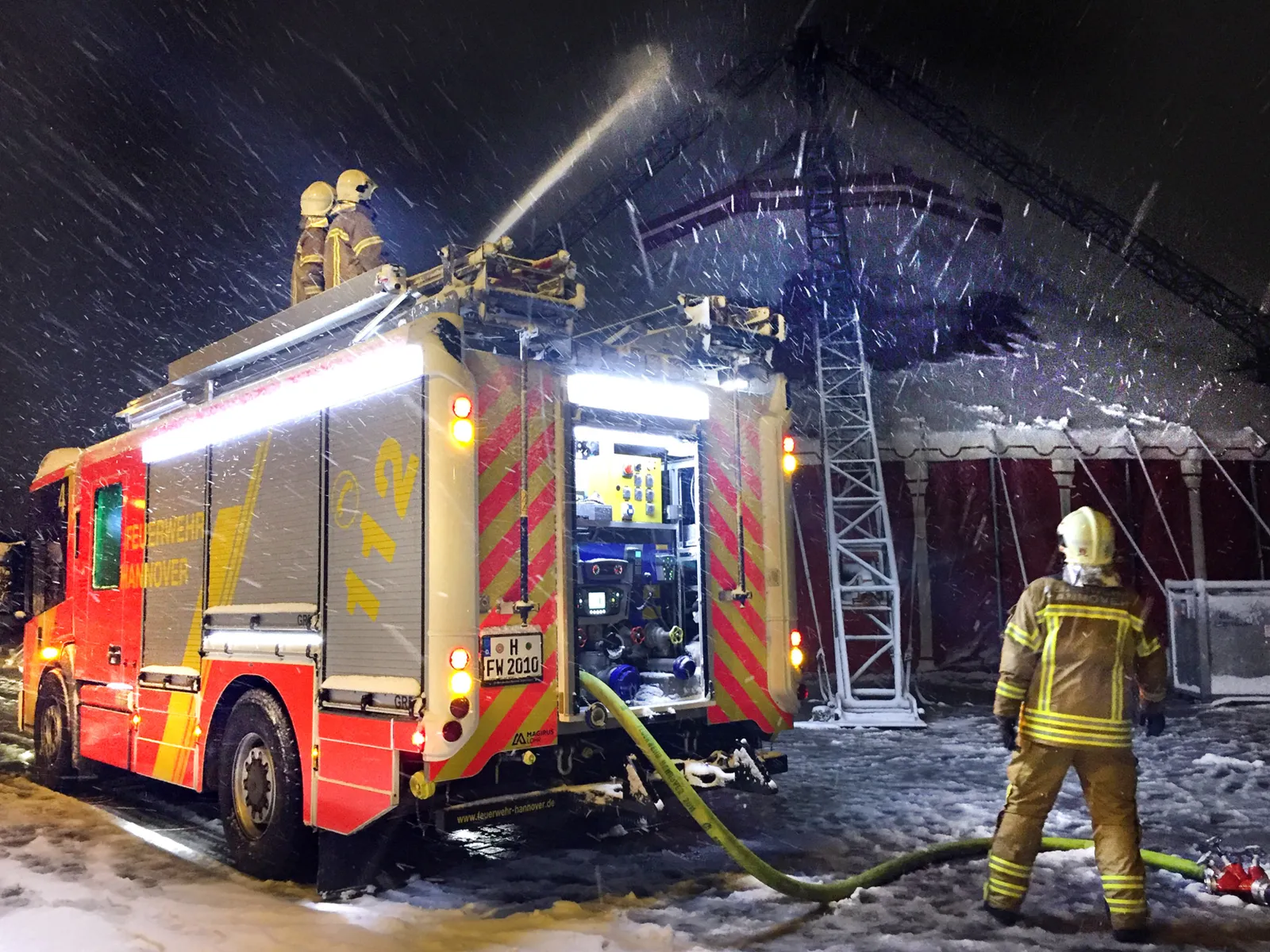 Im Vordergrund ein HLF20 der Feuerwehr Hannover mit mehreren Einsatzkräften, die durch Spülen das Zirkuszelt im Hintergrund von der Schneelast befreien.