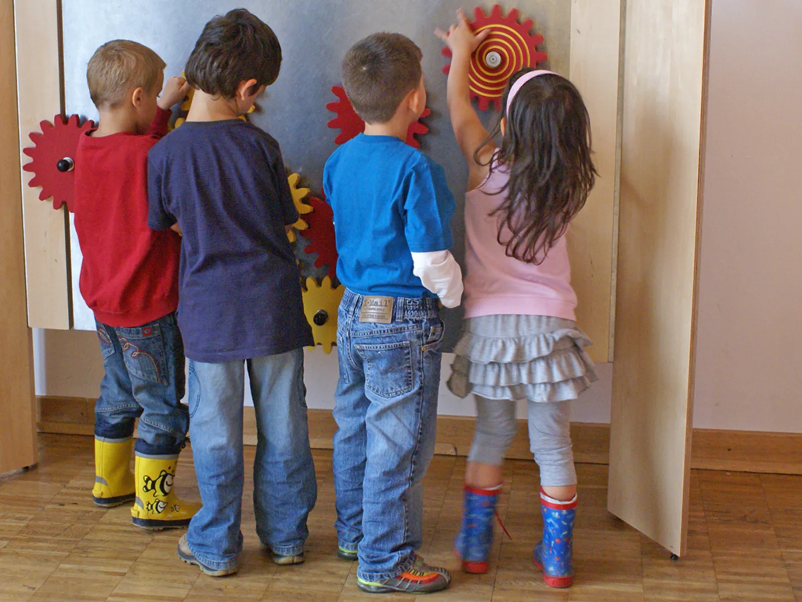 Drei Jungen und ein Mädchen probieren bunt angemalte Holzzahnräder aus, die an einer Ausstellungswand verschraubt sind.