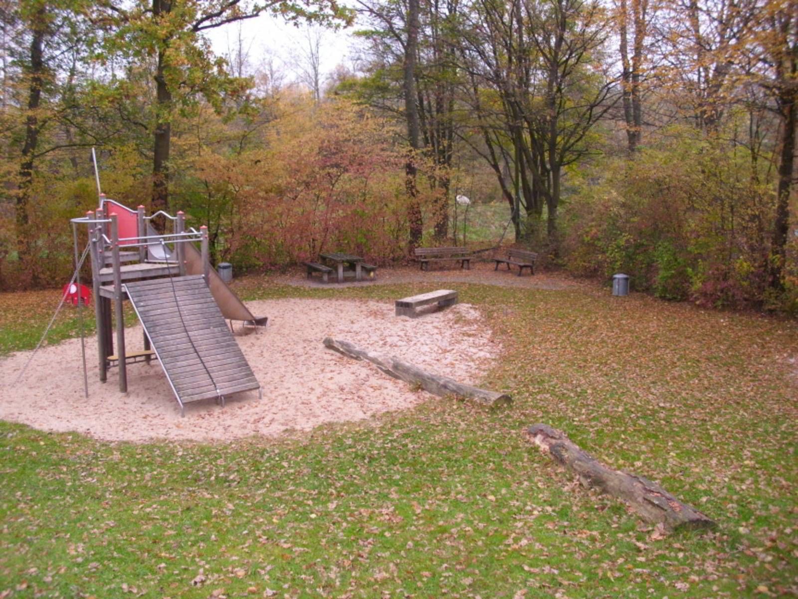 Spielplatz mit Holzrutsche und Sandfläche umrandet von Bäumen und Büschen