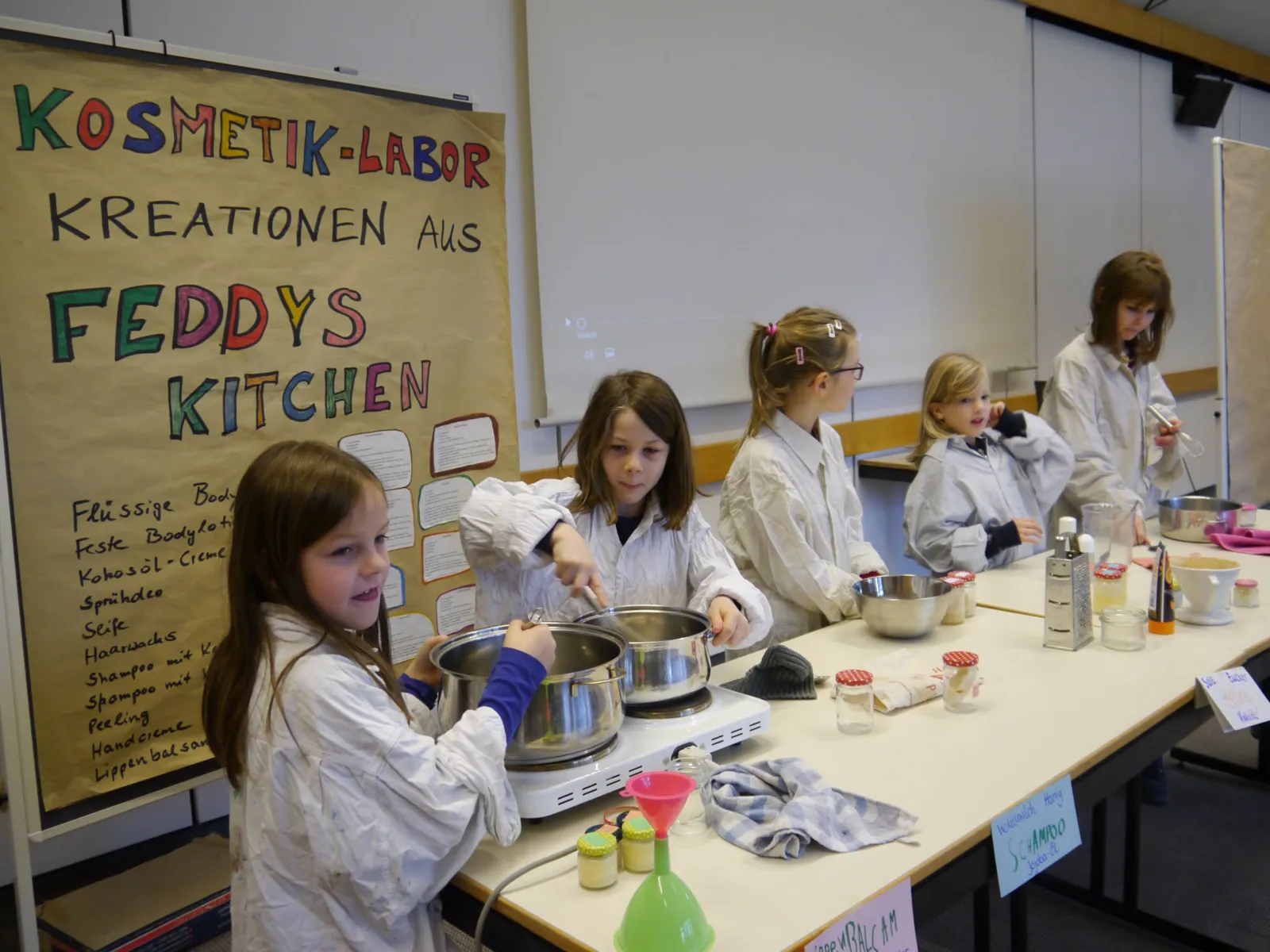 Fünf Kinder im Kittel an Tischen mit Töpfen. 