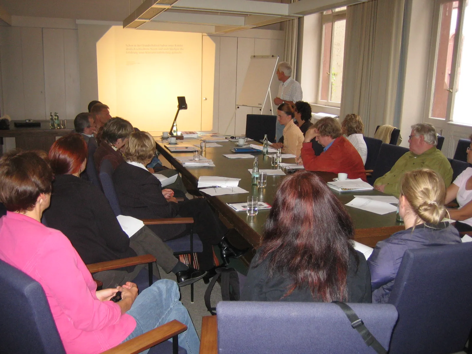 Teilnehmende der Arbeitsgruppe sitzen an einem langen Tisch und hören einen Vortrag