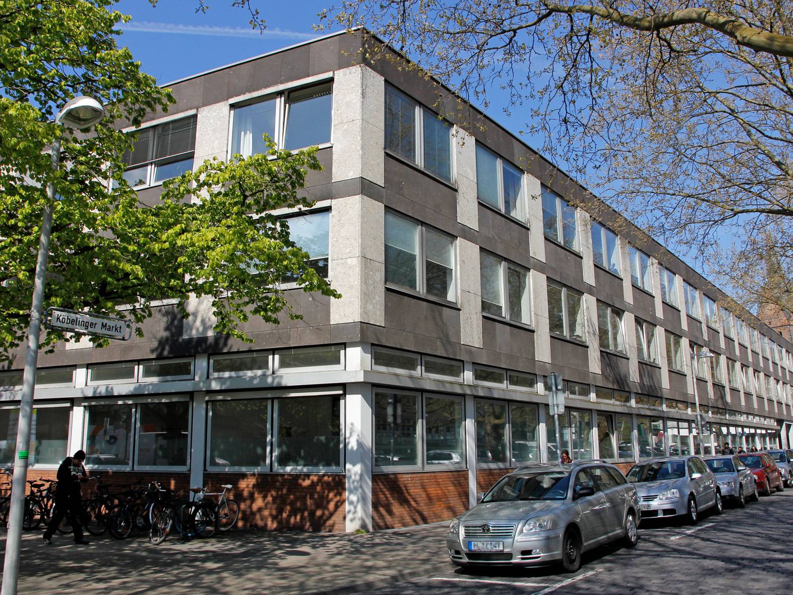 Dreistöckiges Gebäude an der Ecke Leinstraße/Köbelinger Markt mit parkenden Autos und Fahrrädern davor.
