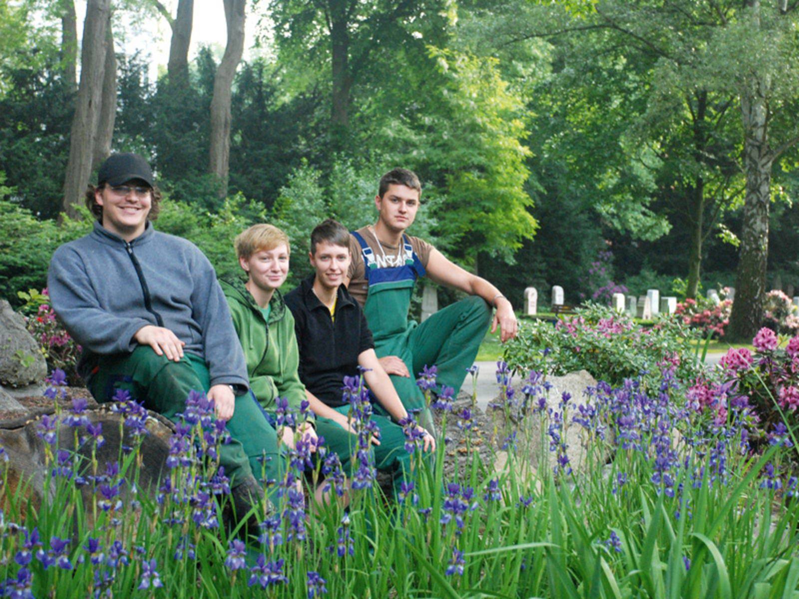 Zwei junge Männer und zwei junge Frauen sitzen in gärtnerischer Arbeitskleidung zwischen blühenden Iris und Rhododendron auf einem städtischen Friedhof