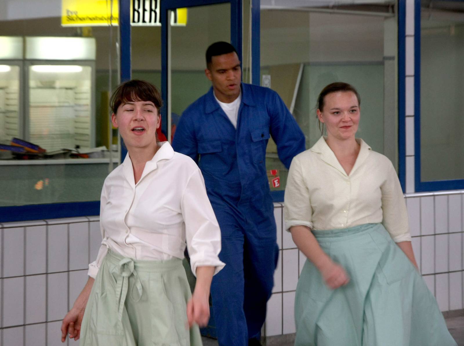 Szene aus einem Stück: Zwei Frauen in türkisen Petticoats kommen aus einer Tankstelle, ihnen folgt ein Mann im Blaumann.