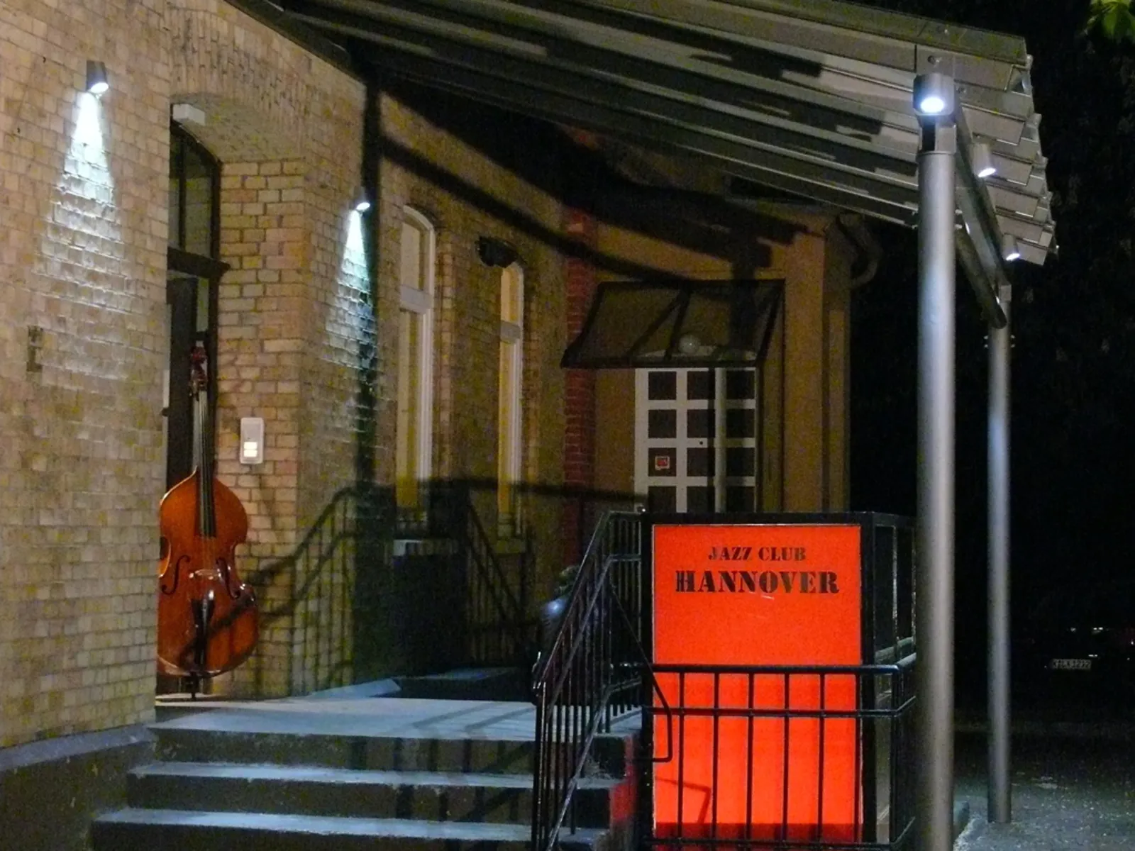 Ein Hauseingang mit überdachter Treppe und einem Schild mit der Aufschrift Jazz Club Hannover, in der geöffneten Tür steht ein Kontrabass.