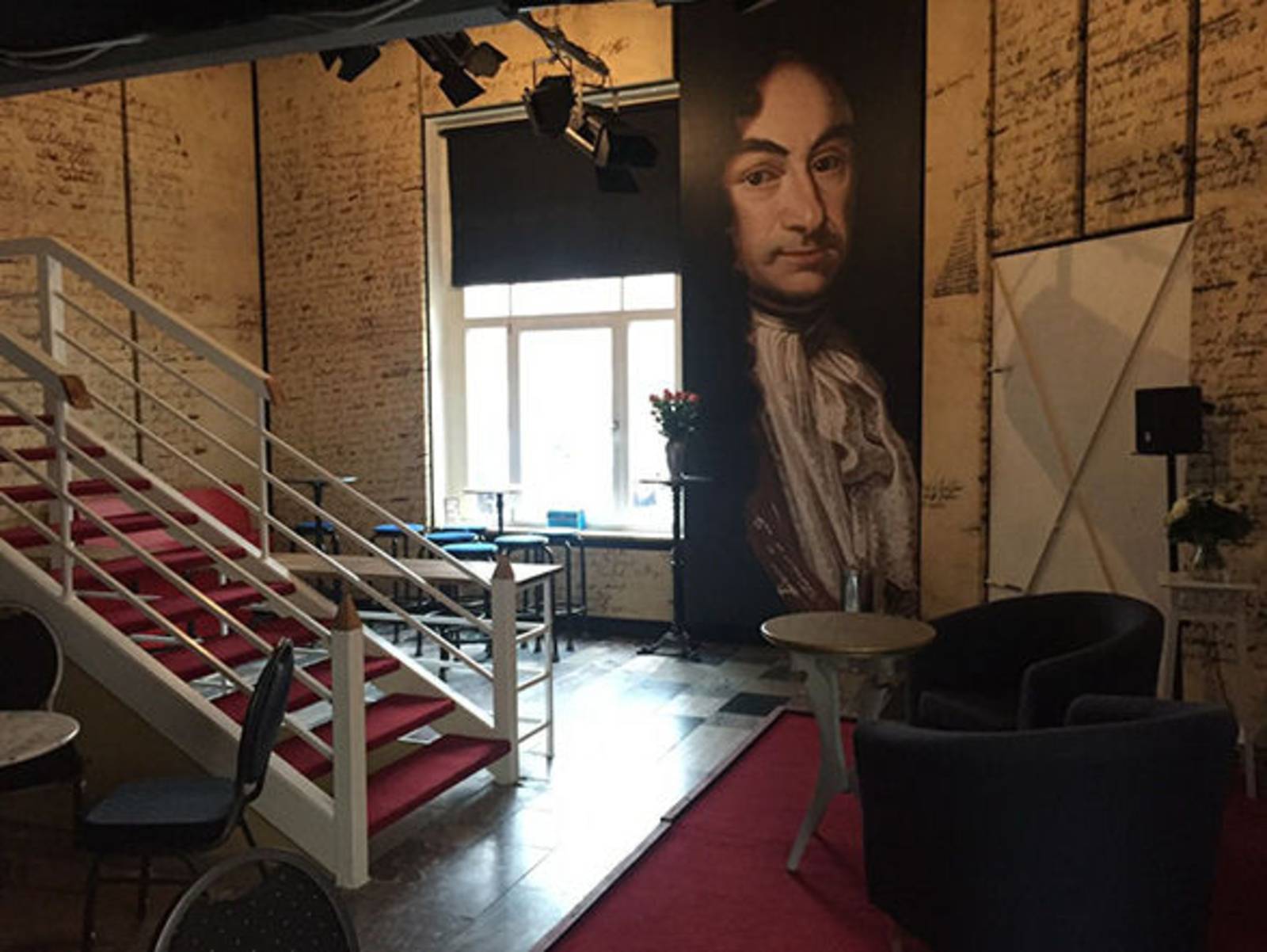 Ein Raum mit Fenster, rotem Teppich, Tischen und Stühlen, einem wandhohen Bild von Leibniz und einer weißen Treppe nach oben,