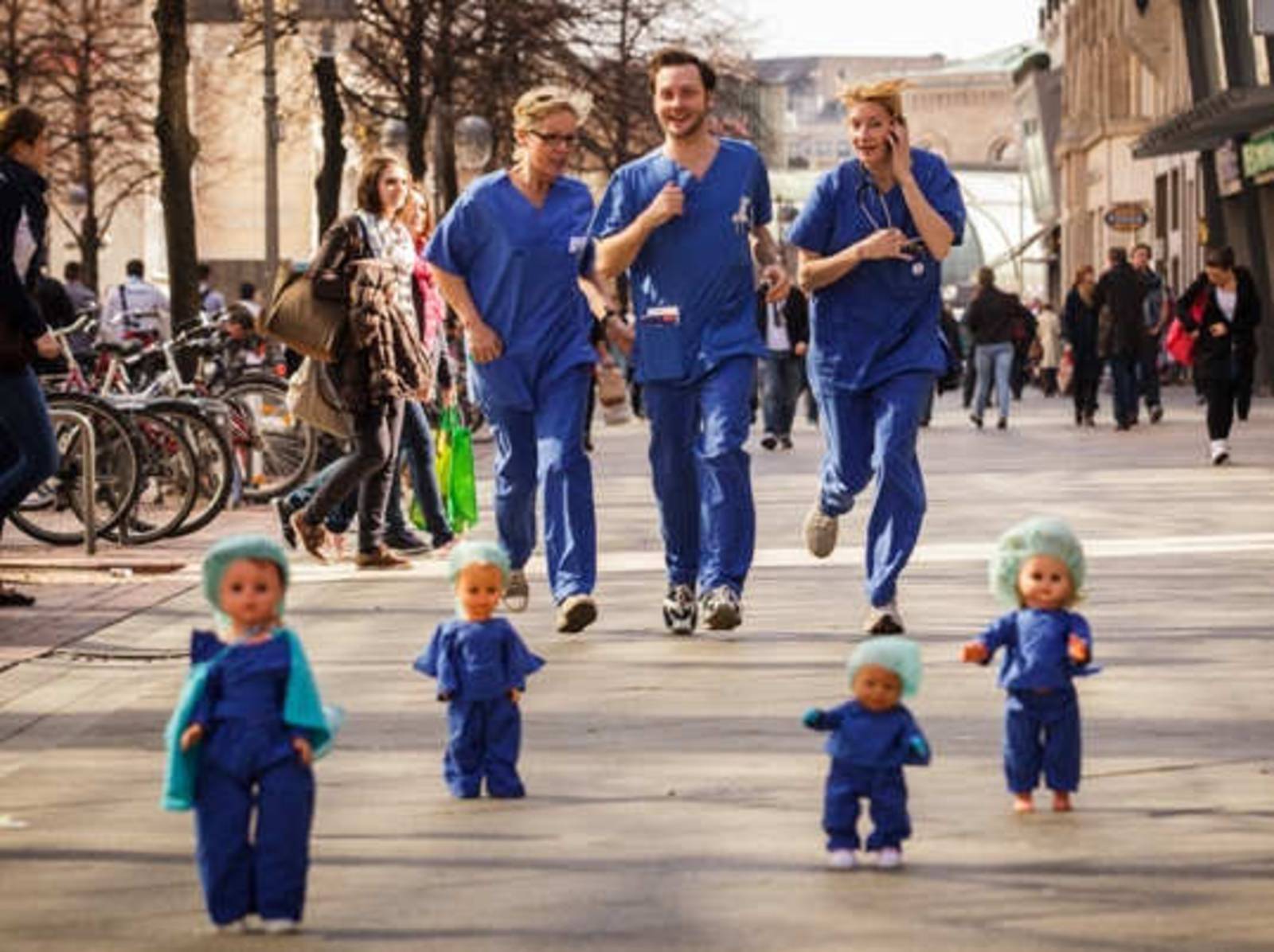Zwei Frauen und ein Mann in blauer OP-Kleidung laufen durch die hannoversche Fußgängerzone. Sie sind umringt von stehenden Kinderpuppen, die genauso gekleidet sind wie sie.