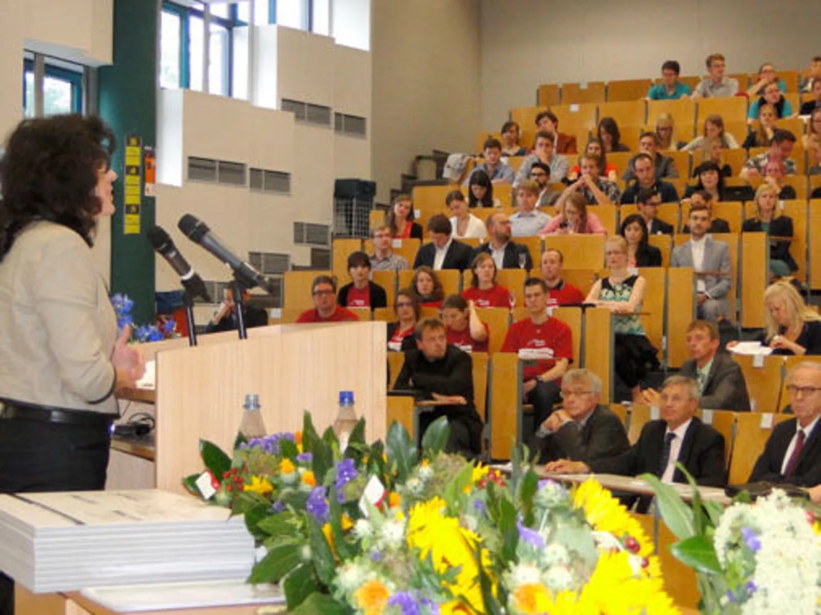 Viele Menschen sitzen in einem Hörsaal, eine Rednerin steht an einem Pult.